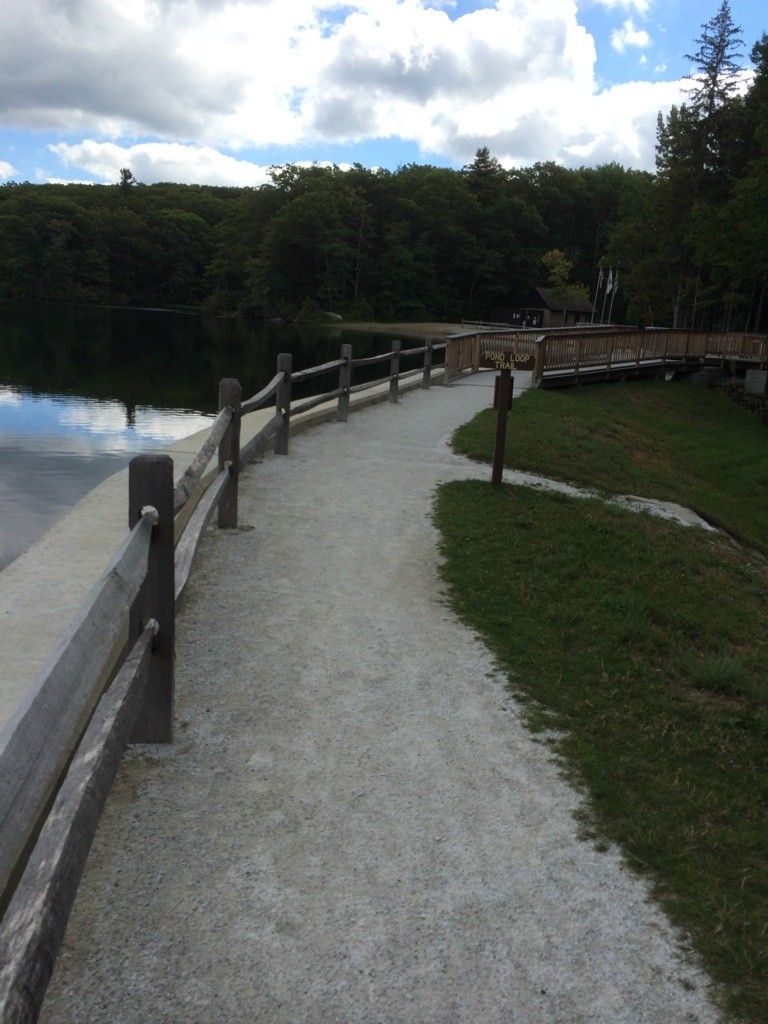 Paved path with wooden railing beside a lake under a cloudy sky. - Benedict Pond Loop Trail