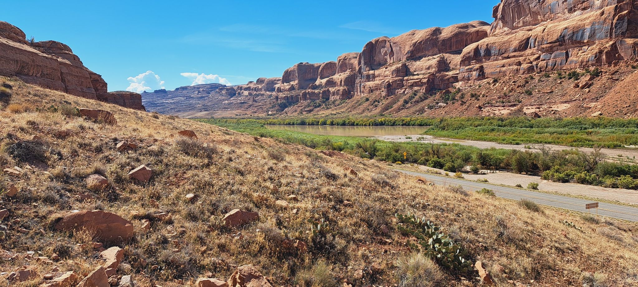 Wide desert canyon with a river, red cliffs, and dry brush under blue sky. - Corona Arch Trail