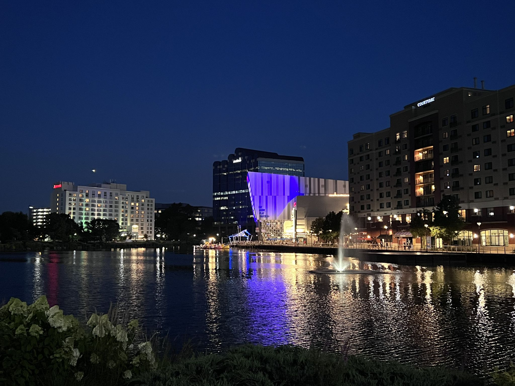 Nighttime urban waterfront with illuminated buildings reflecting on dark water and fountain. - Rio Boardwalk