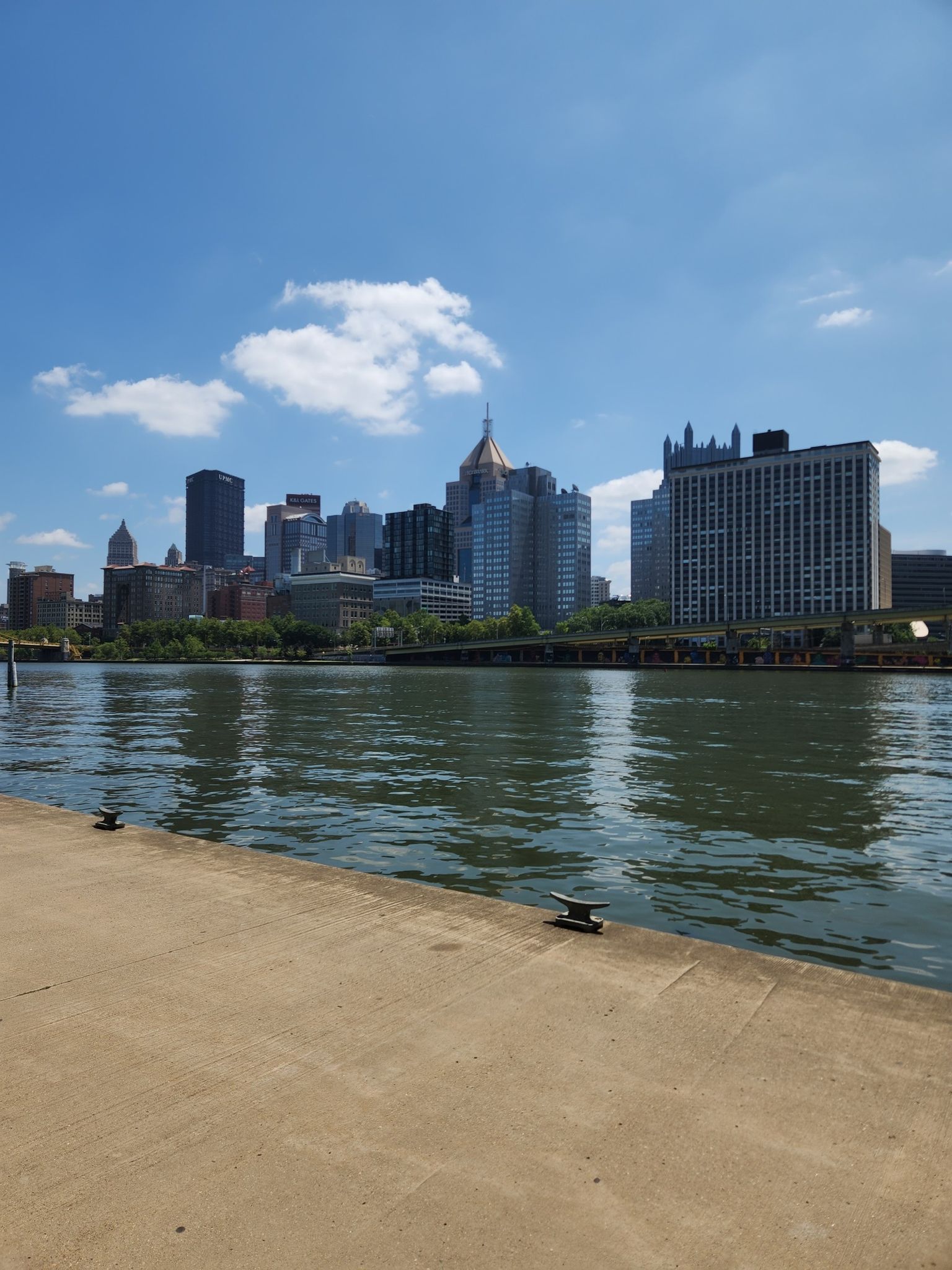 City skyline across a river from a concrete dock under a bright blue sky. - Three Rivers Heritage Trail