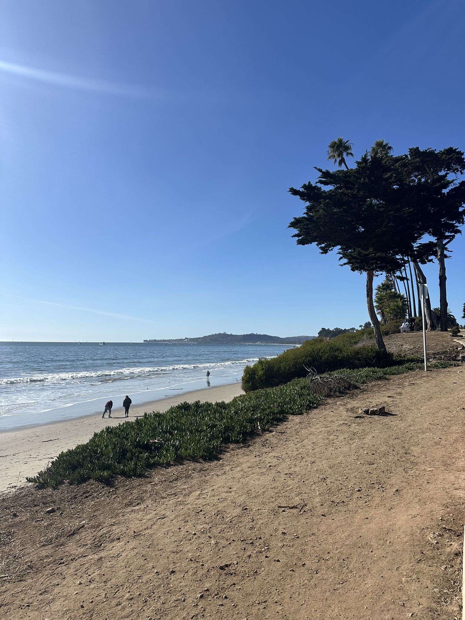 Bright sunny beach with people walking, ocean waves, and large trees. - Butterfly Beach Santa Barbara – Butterfly Beach loop from Montecito Inn