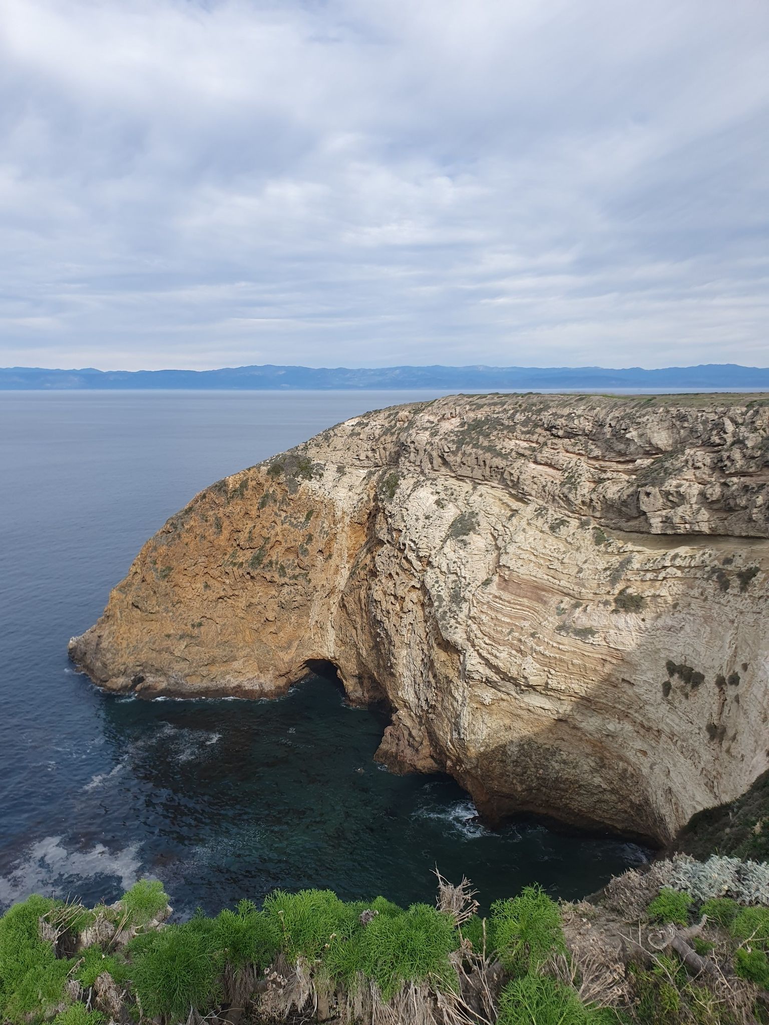 Layered sea cliff with a large cave, calm ocean, and cloudy sky. - North Bluff Trail