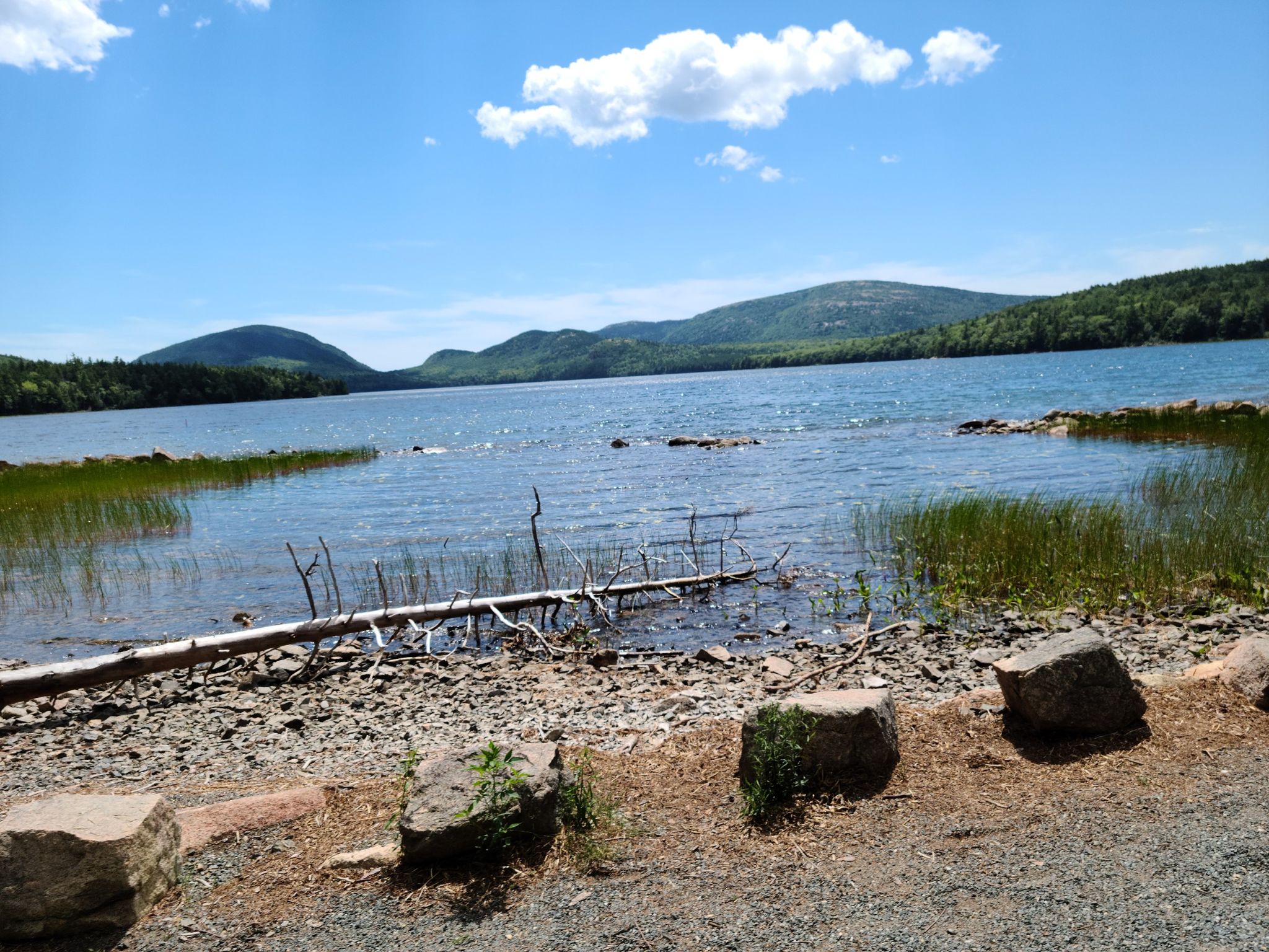 Calm lake with rocky shore, distant mountains, and blue sky with white clouds. - Eagle Lake Loop