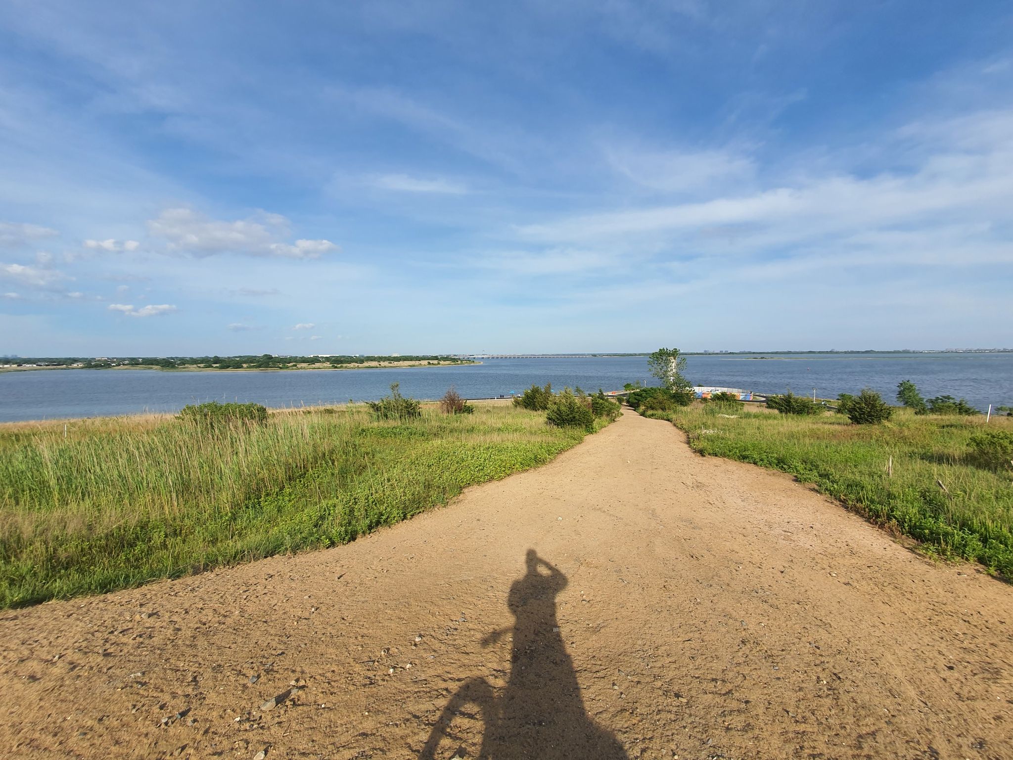 Shadow of a cyclist on a dirt path leading to a sunny bay. - Great Blue Heron Loop