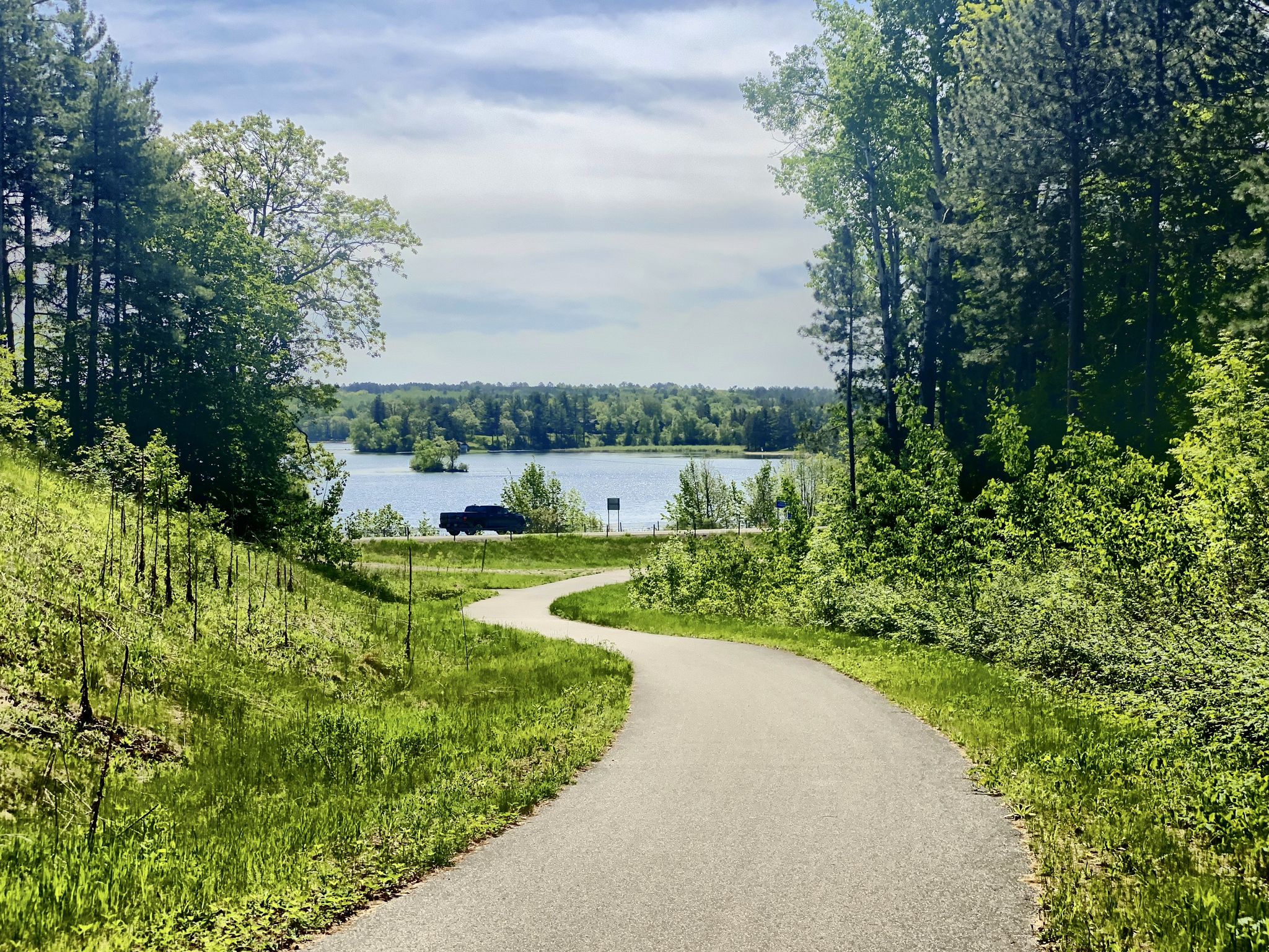 Winding paved path leading to a lake with a car, surrounded by green trees. - Paul Bunyan and Heartland Trail Loop