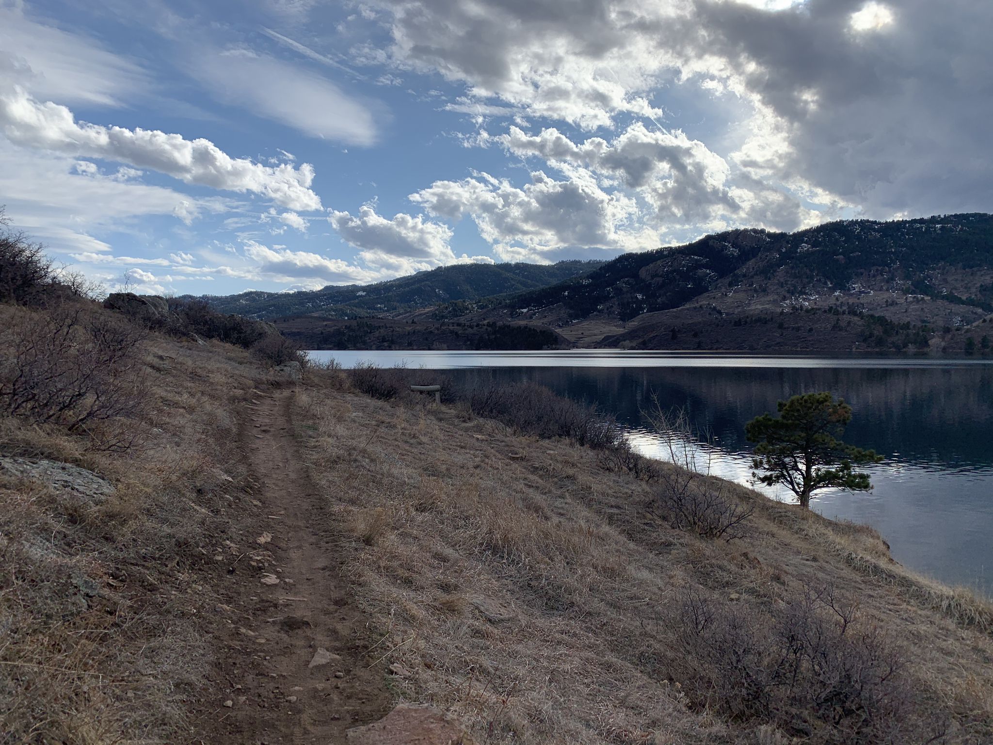 Dirt path on dry hillside overlooking a calm lake and mountains under cloudy sky. - Sunrise Beach – Horsetooth Reservoir loop from Saddle Ridge Natural Area
