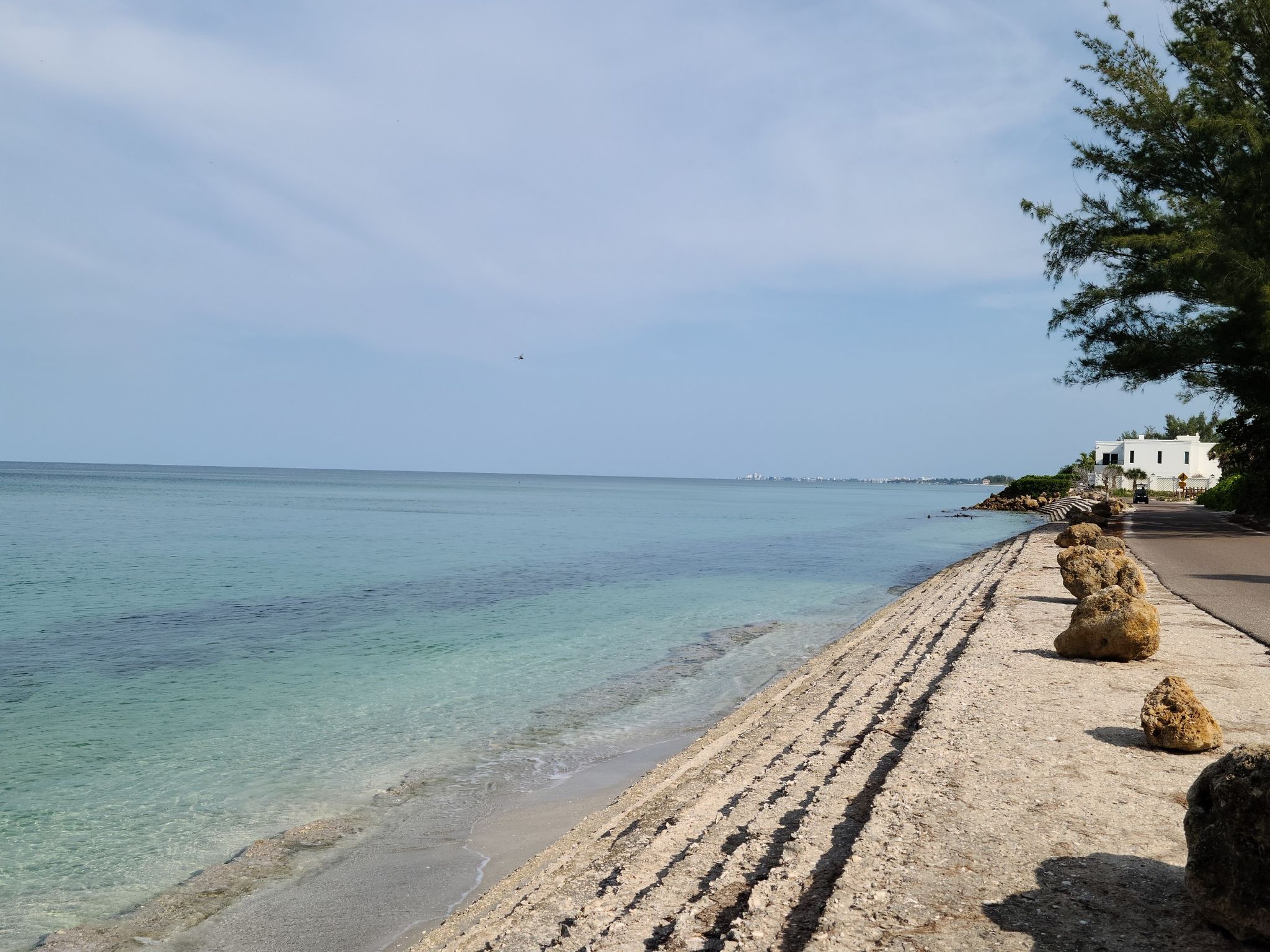 Bright sunny day over clear turquoise ocean next to a rocky shore and road. - Casey Key Road Scenic Drive