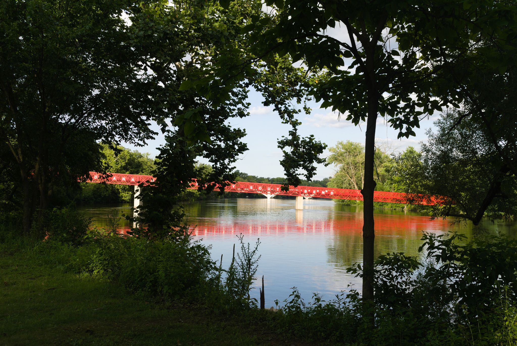 A vibrant red bridge spans a river, framed by dark green trees. - St Joseph River bridge loop from South Bend