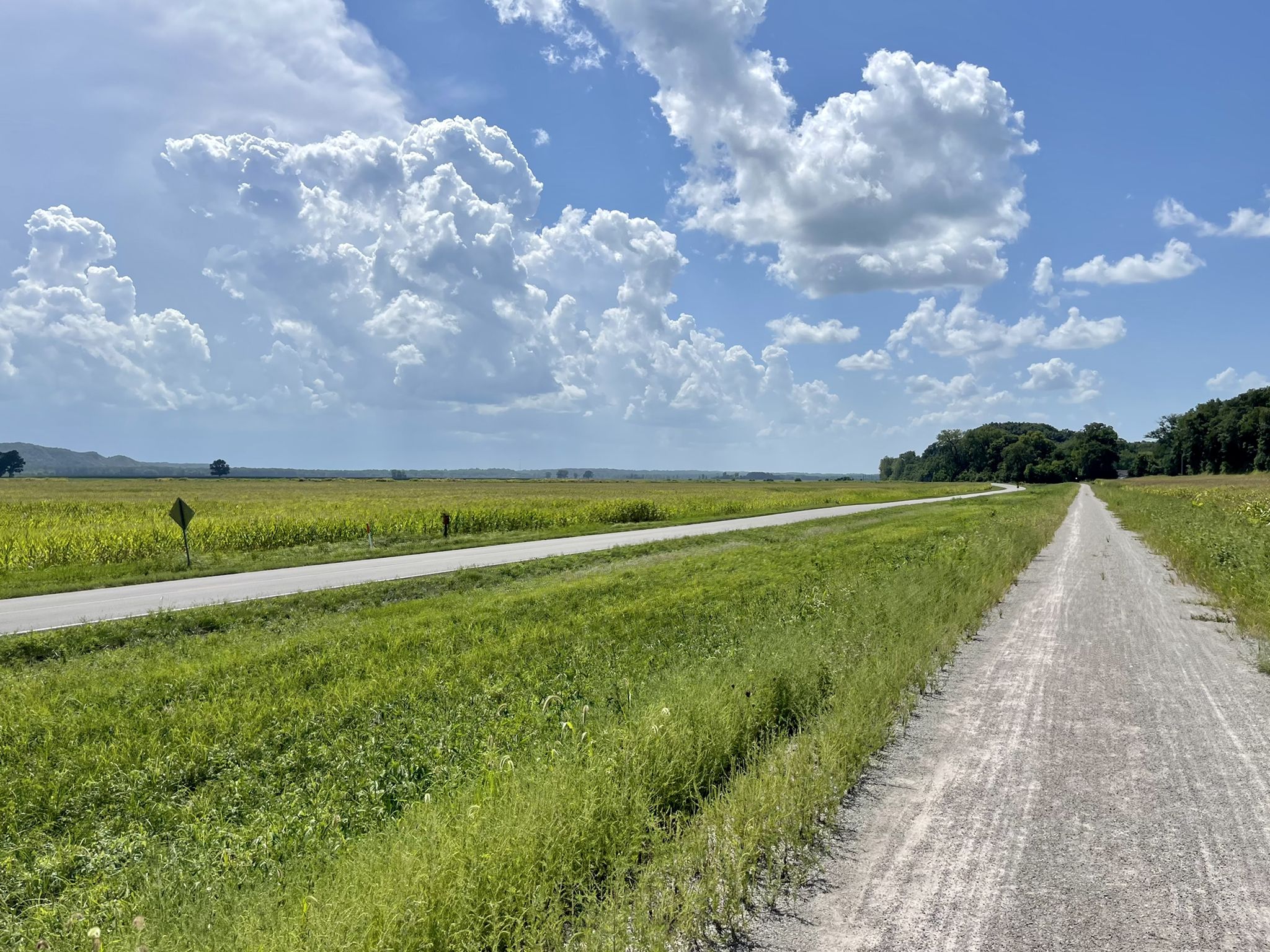Dirt path and paved road beside a vast green field under a cloudy sky. - Katy Trail: Jefferson City to Hermann