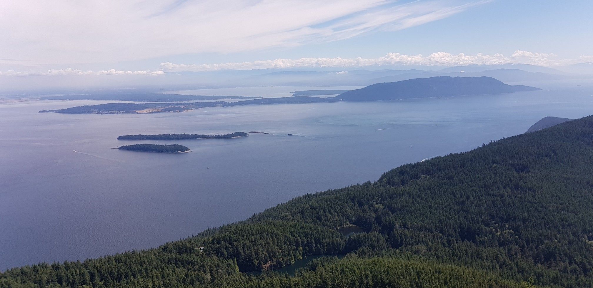Aerial view of a forested coastline, vast water, and distant islands under a hazy sky. - Cascade Falls – Mount Constitution loop from Orcas Island