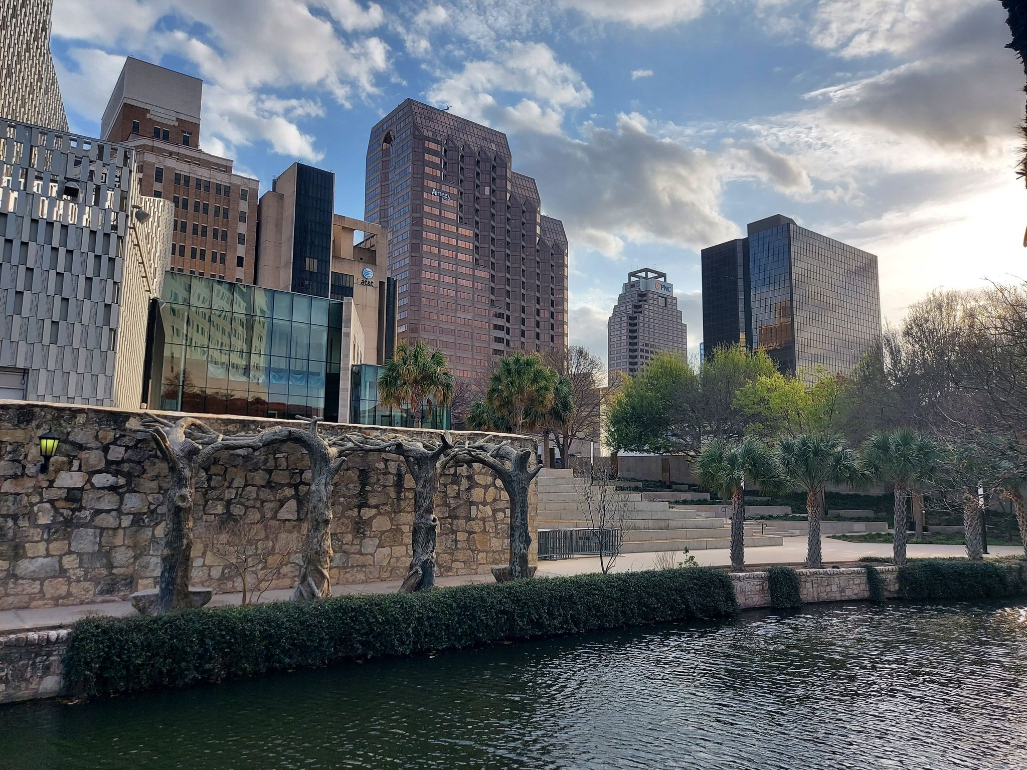 River with stone archway, tall city buildings, and palm trees at sunset. - Alamo Mills Dam – River Walk loop from San Antonio