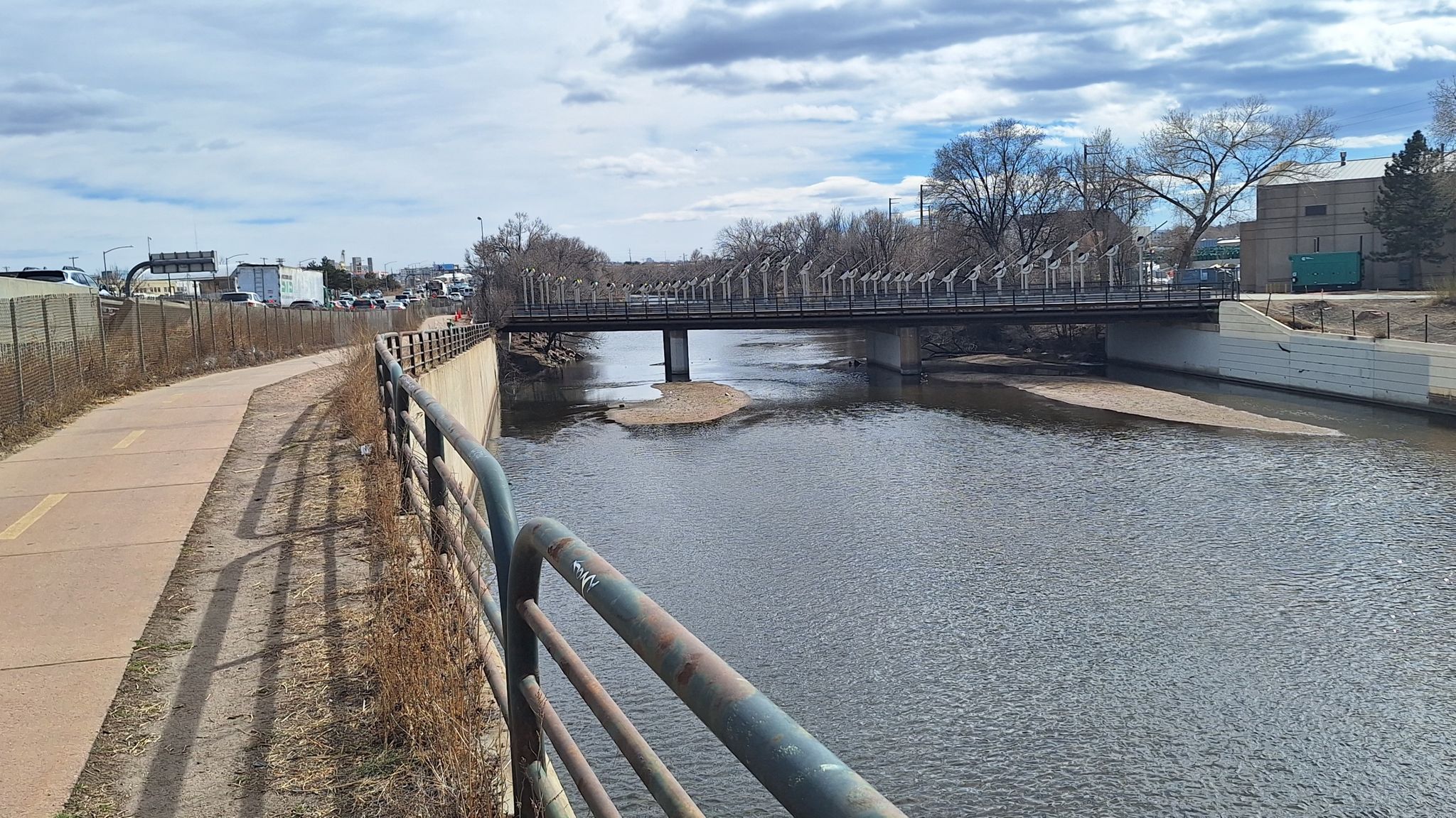 Paved path alongside a river with a bridge and industrial buildings under a cloudy sky. - South Platte River Trail
