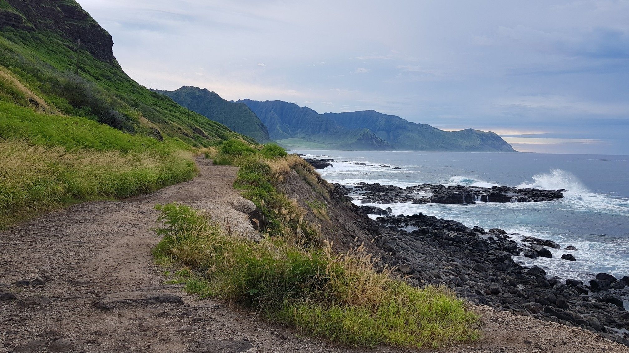 Dirt path alongside a rocky coastline with green mountains and crashing waves. - Sea arch at Kaʻena Point – Yokohama Bay loop from Kaena Point Satellite Tracking Station