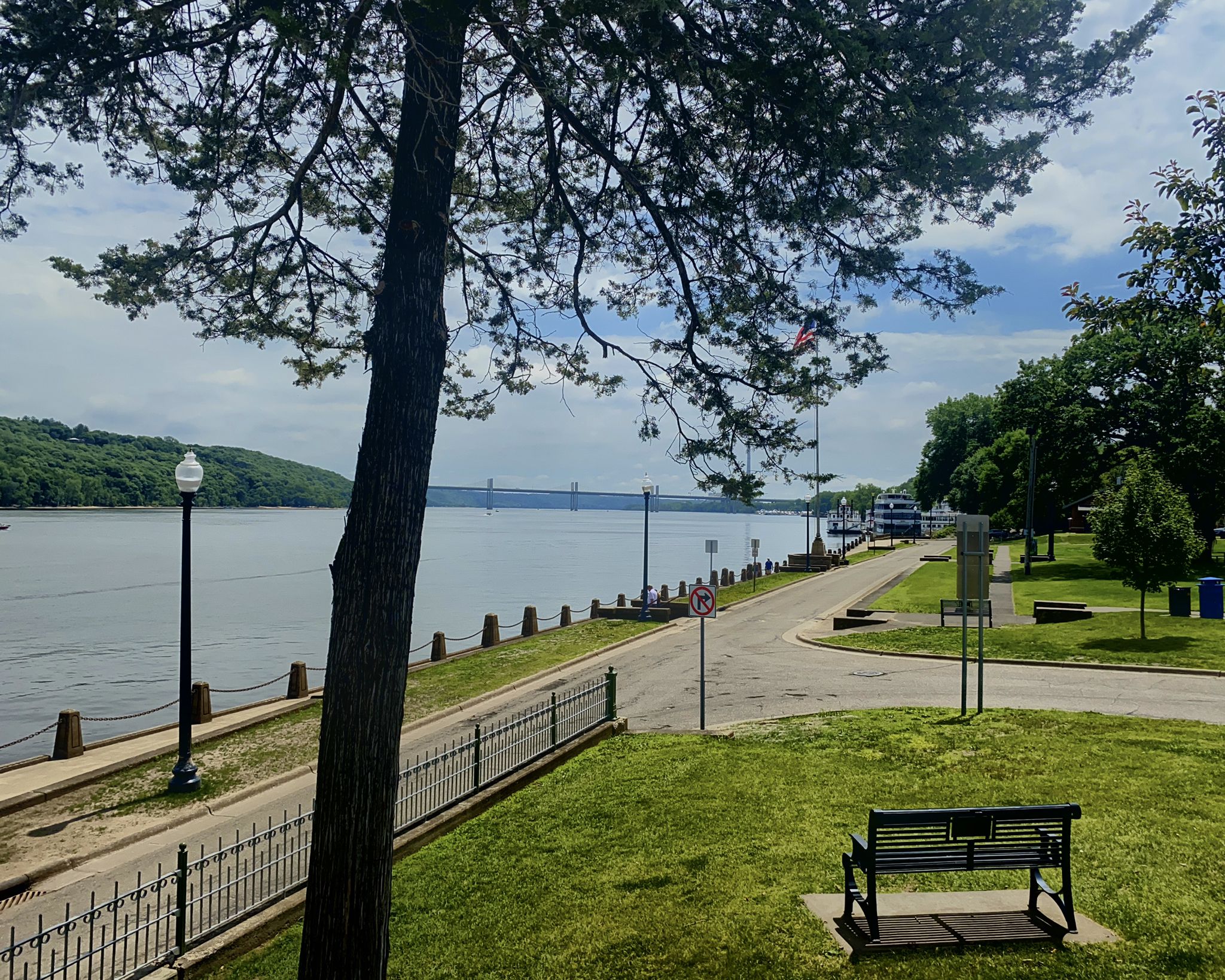 A park bench under a large tree overlooks a riverfront path and distant bridge. - Stillwater Lift Bridge – Stillwater Lift Bridge loop from Stillwater