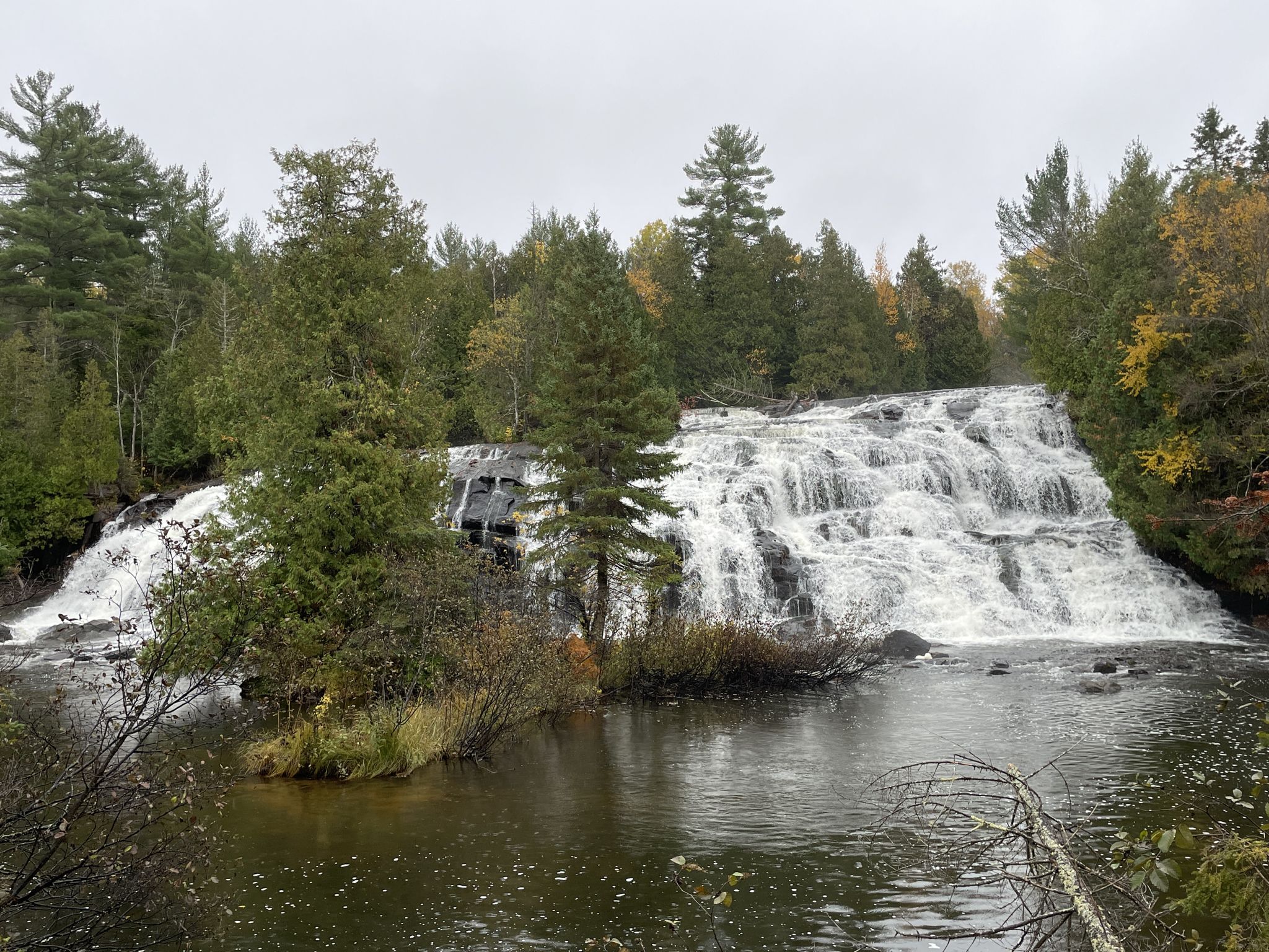 Multi-tiered waterfall cascades into a river, surrounded by autumn trees under an overcast sky. - Bond Falls Loop