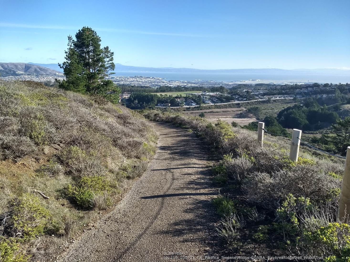Dirt path winds through sunny hills overlooking a distant town and ocean. - Notch Trail Overlook – Nike Missile Site loop from Milagra Ridge