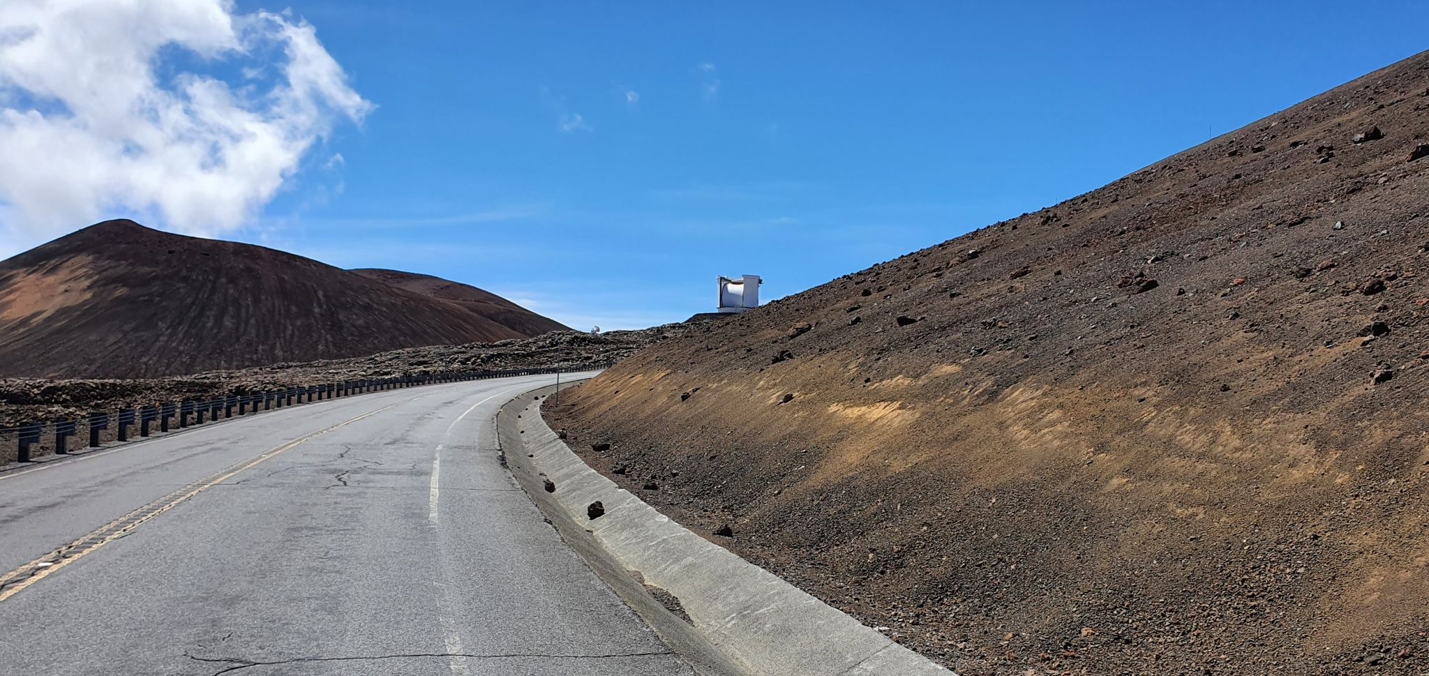 A paved road winds uphill past barren slopes under a clear blue sky. - Mauna Kea summit from Puʻu Huluhulu