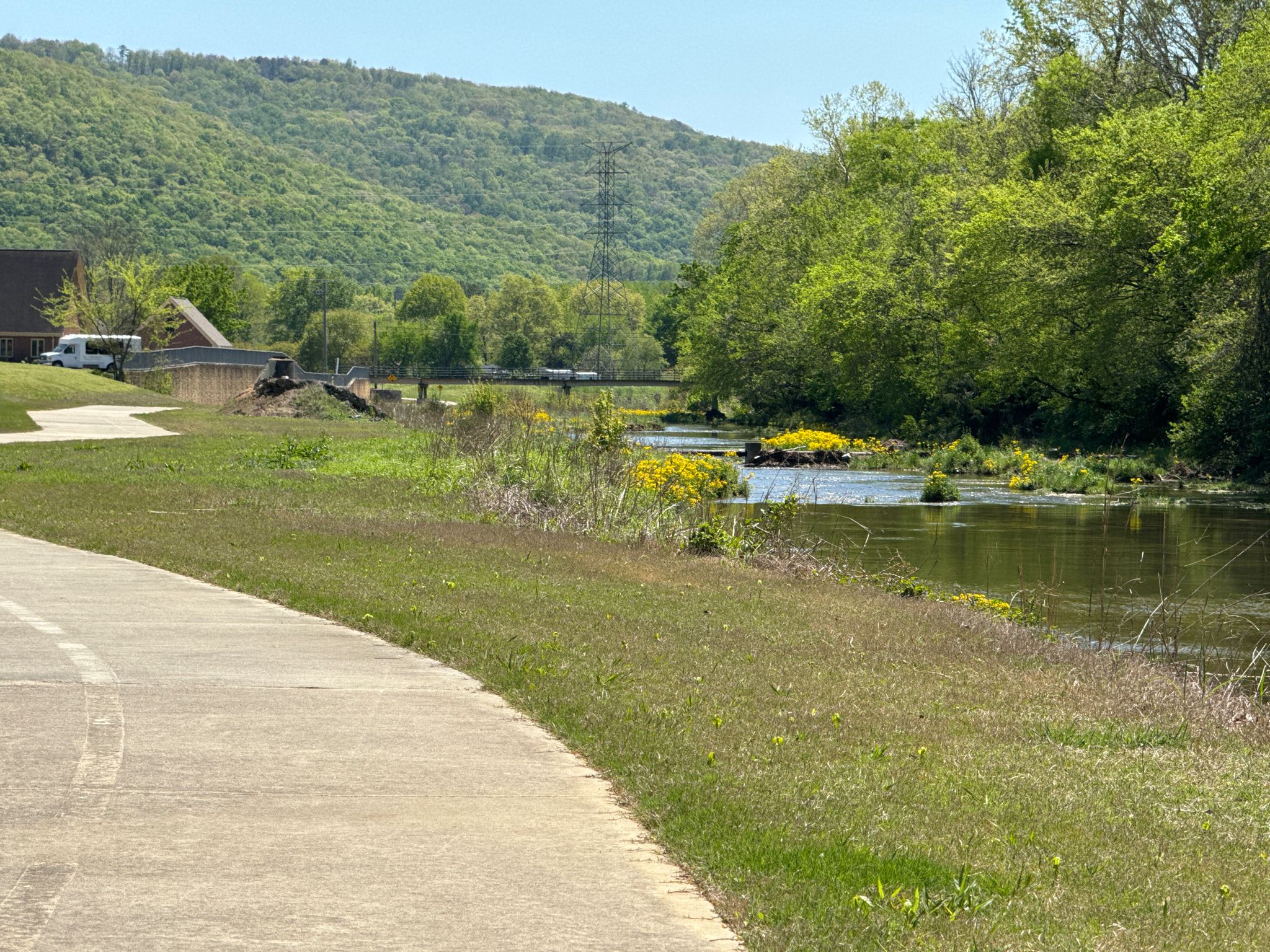 A paved path curves beside a river with yellow flowers, green trees, and hills. - Aldridge Creek Greenway – Aldridge Creek Greenway loop from Huntsville
