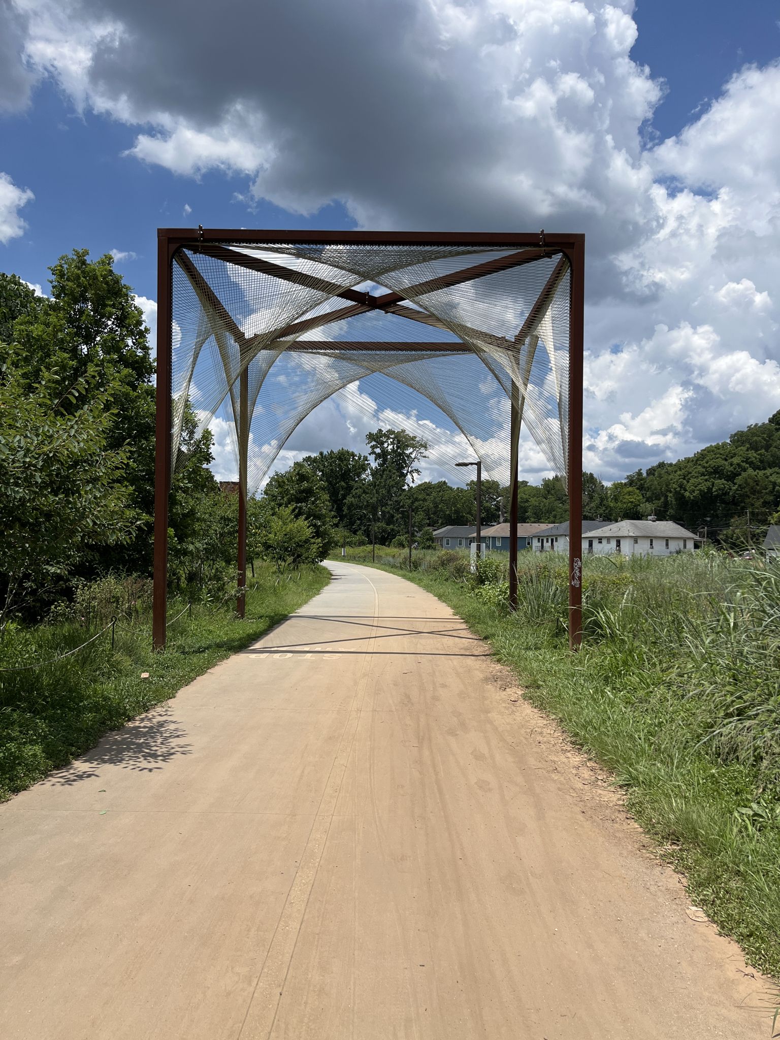 A dirt path leads to a large, open metal archway under a cloudy sky. - Atlanta Beltline Westside Trail