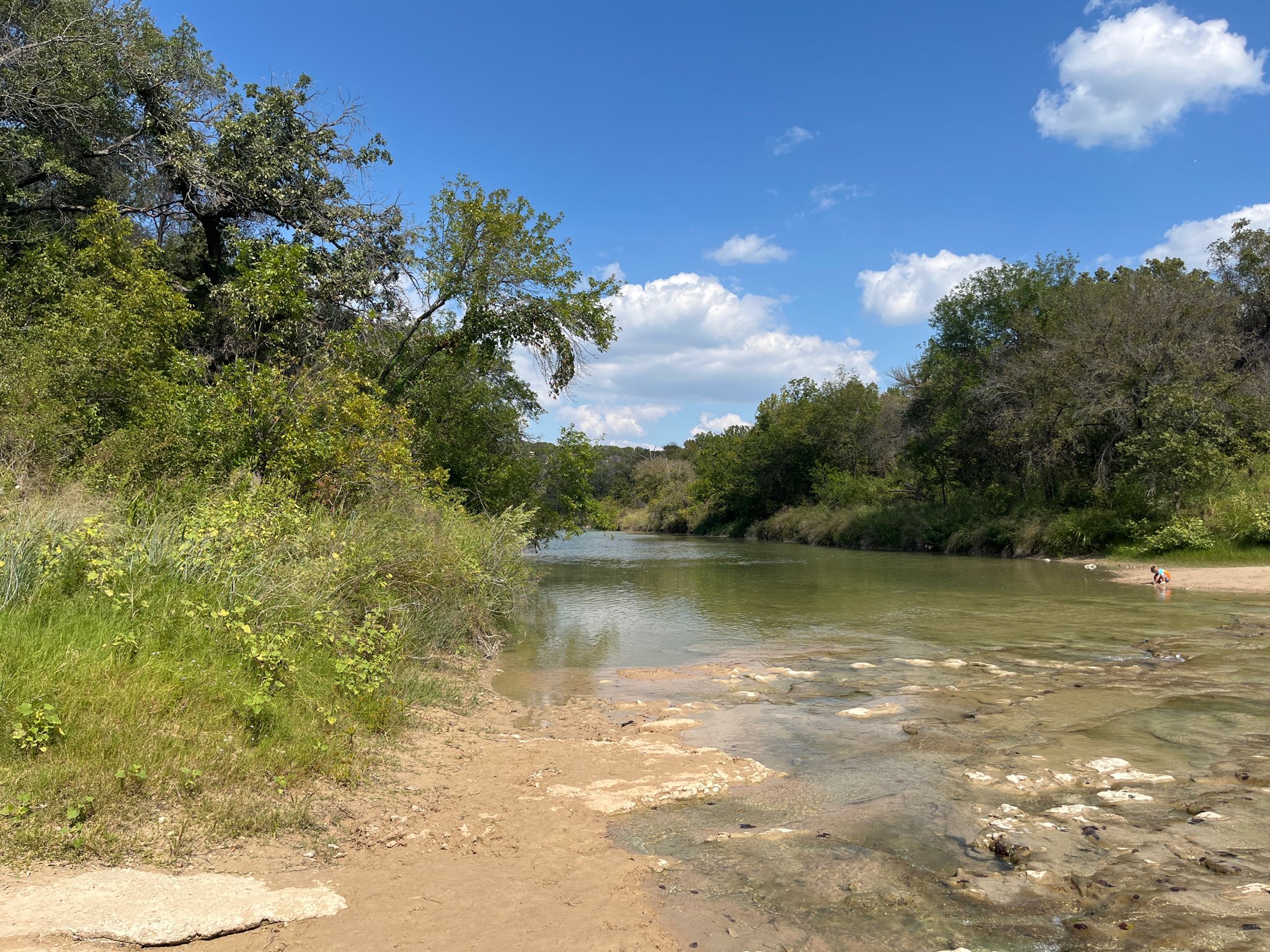 A wide river with a sandy bank and lush green trees under a blue sky. - Dinosaur Valley State Park loop from Dinosaur Valley State Park
