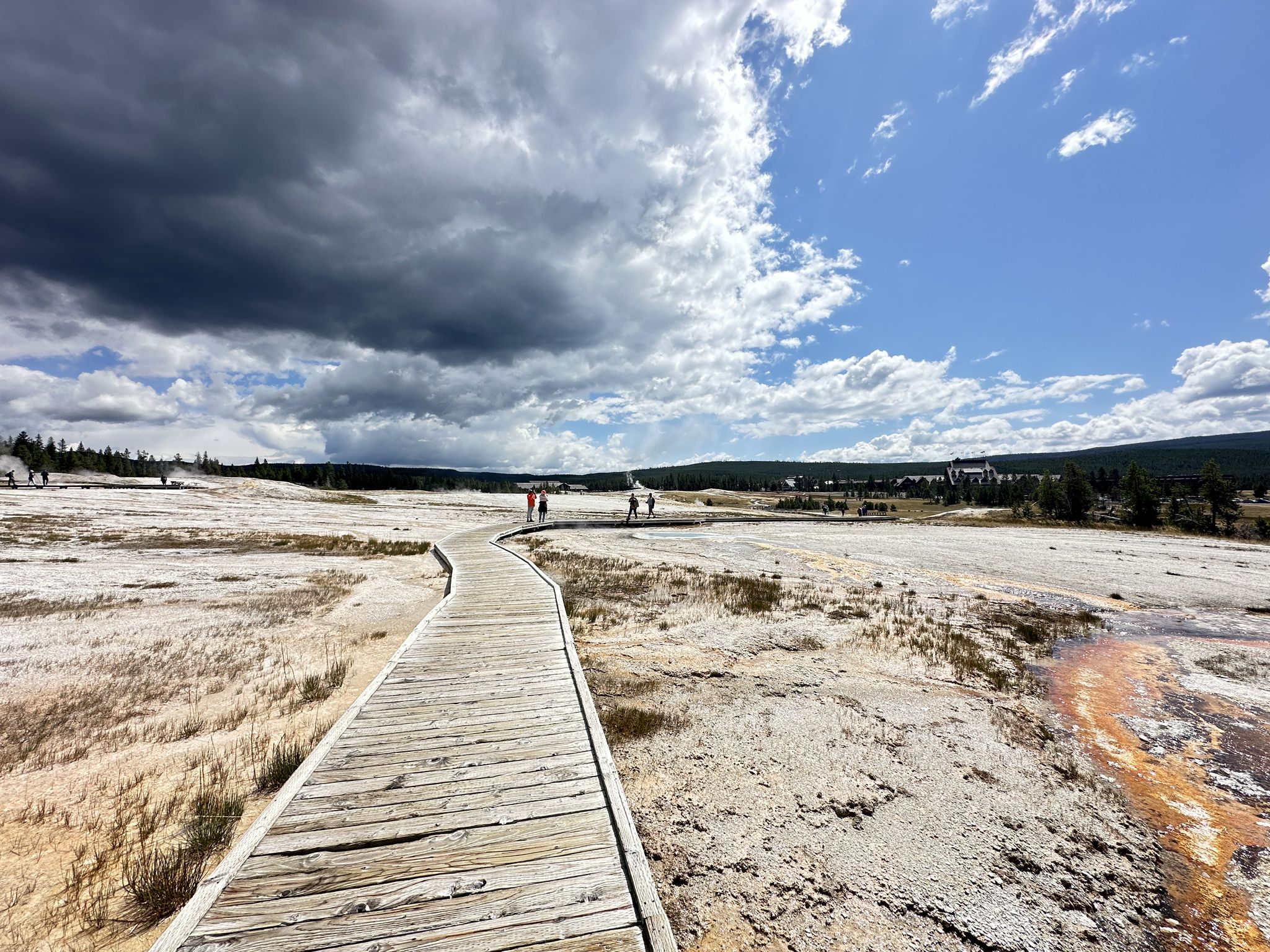 Wooden boardwalk across a barren, steaming landscape with distant people. - Old Faithful Geyser – Blue Star Spring loop from Old Faithful Inn