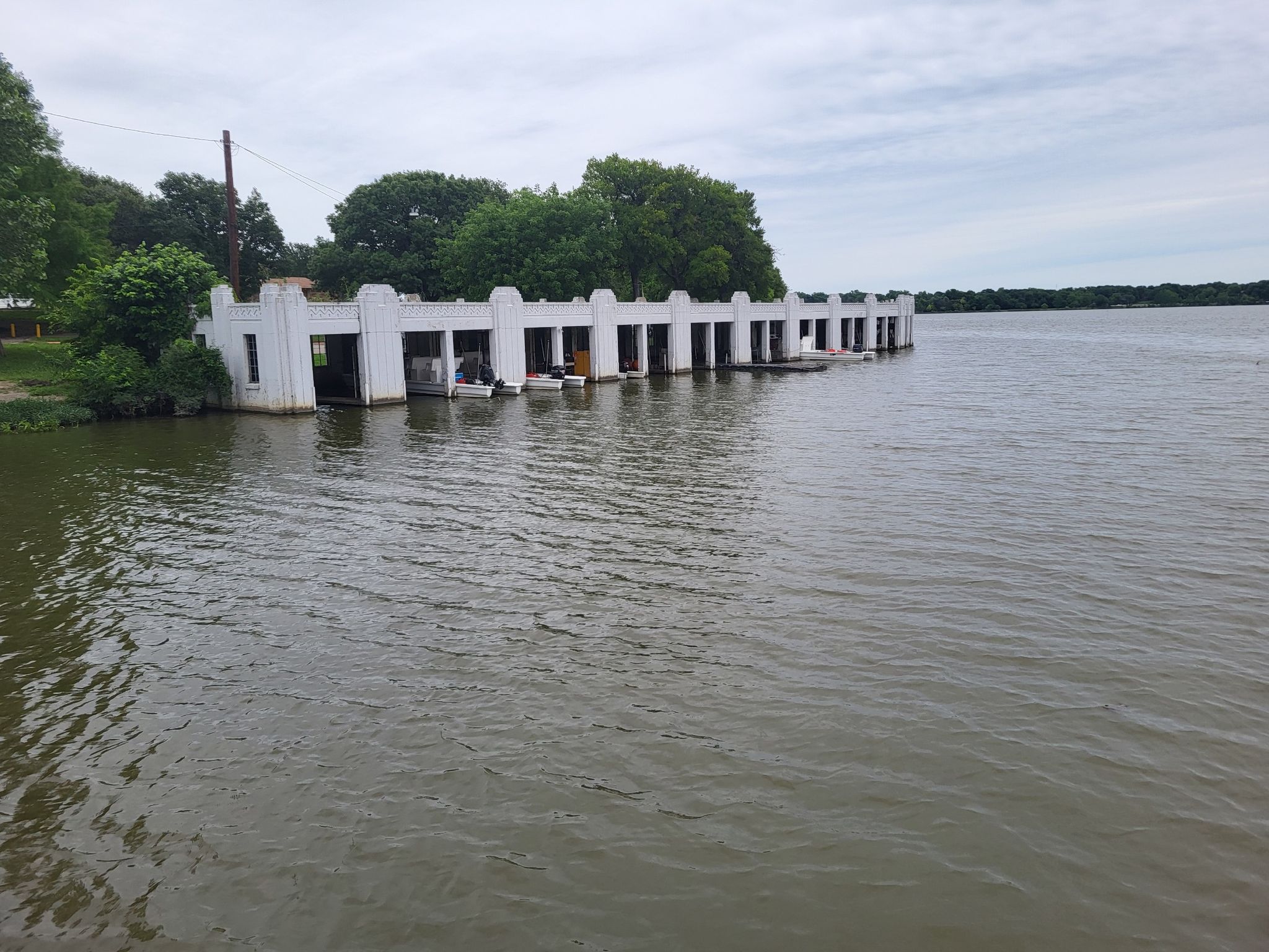 A long white boathouse structure extends into a wide lake under a cloudy sky. - SoPac Trail – Pier at White Rock Lake loop from Park Lane