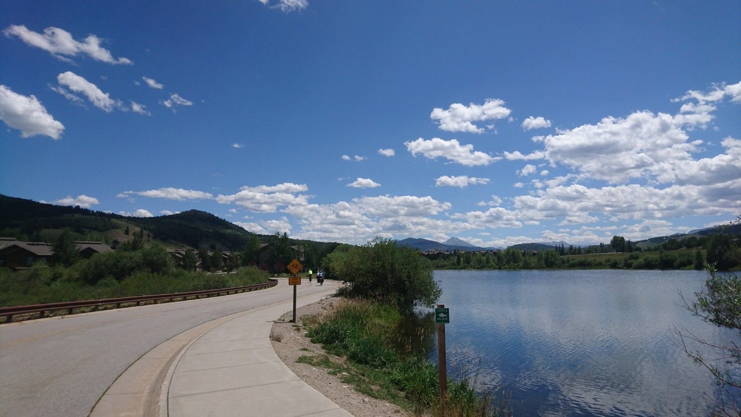 Sunny day with a paved path beside a calm lake and distant hills. - North Pond Park loop from Willowbrook