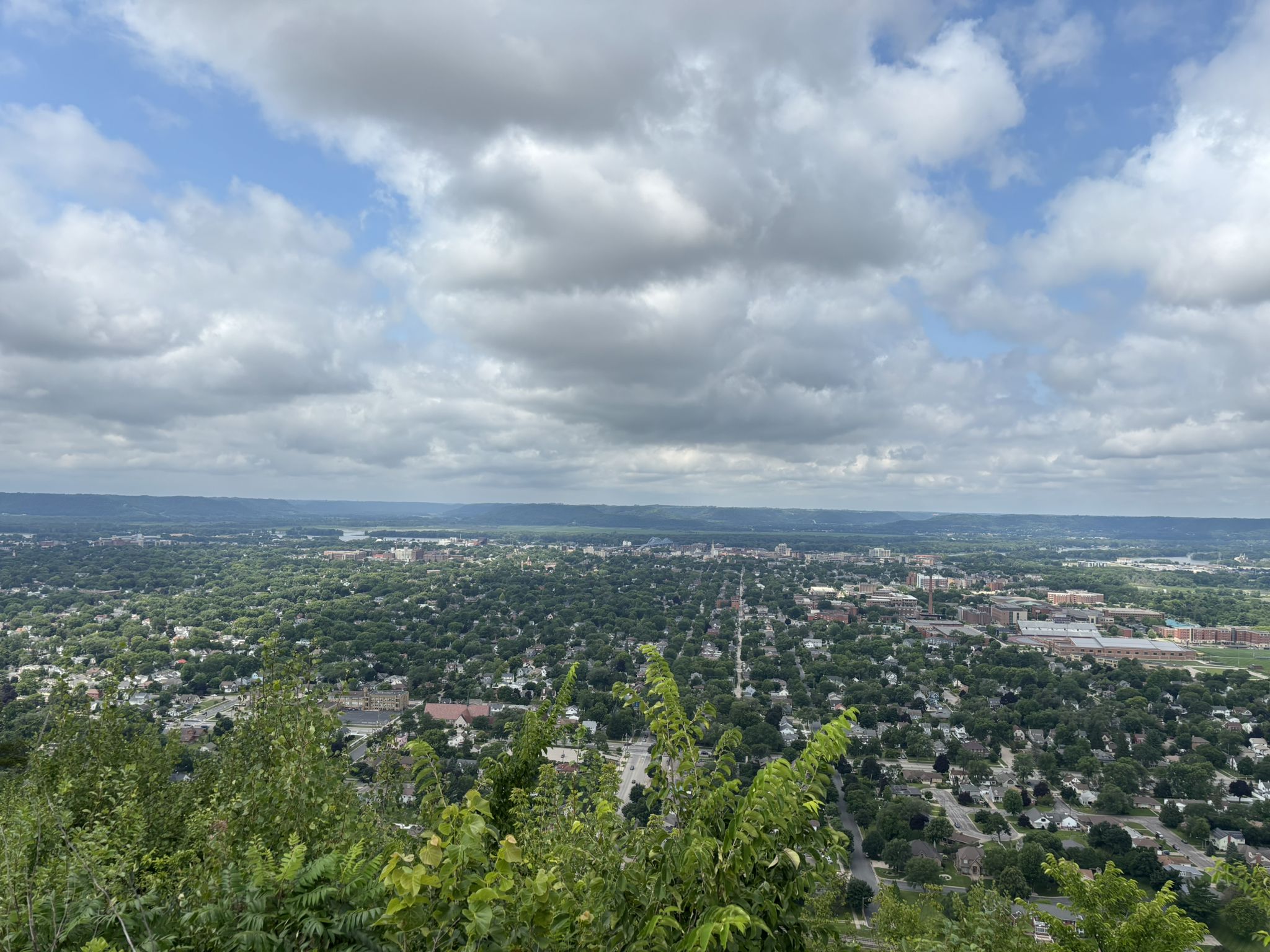 High-angle view of a sprawling town surrounded by green trees under cloudy sky. - Compass Loop