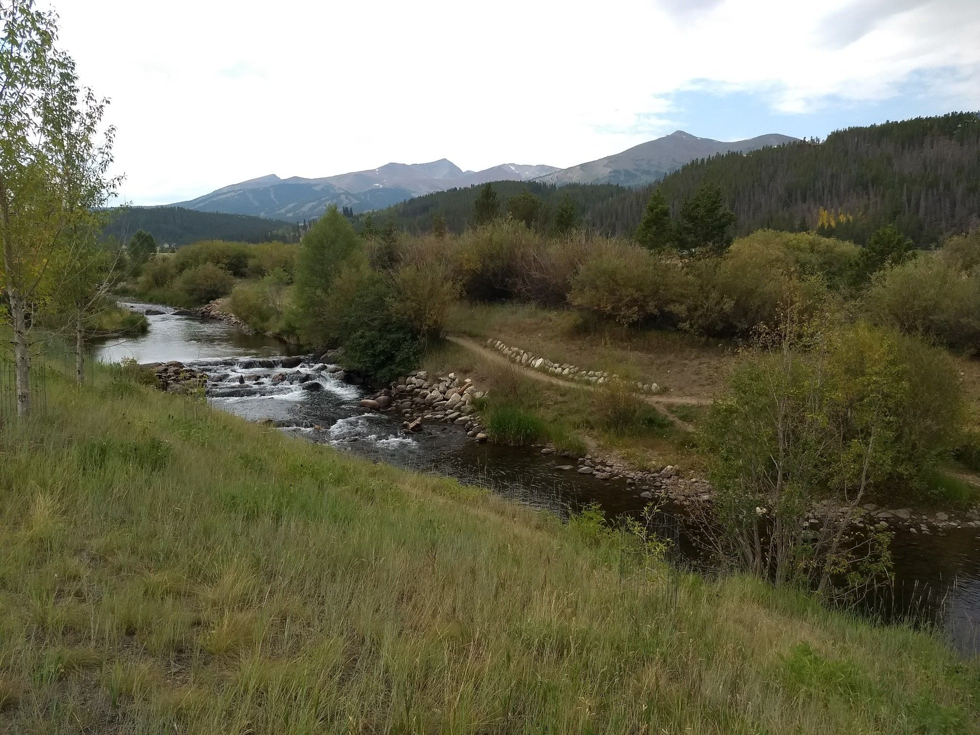 A river flows over rocks, with green banks and distant mountains under clouds. - Breckenridge – Breckenridge Peak 6 condos loop from Breckenridge