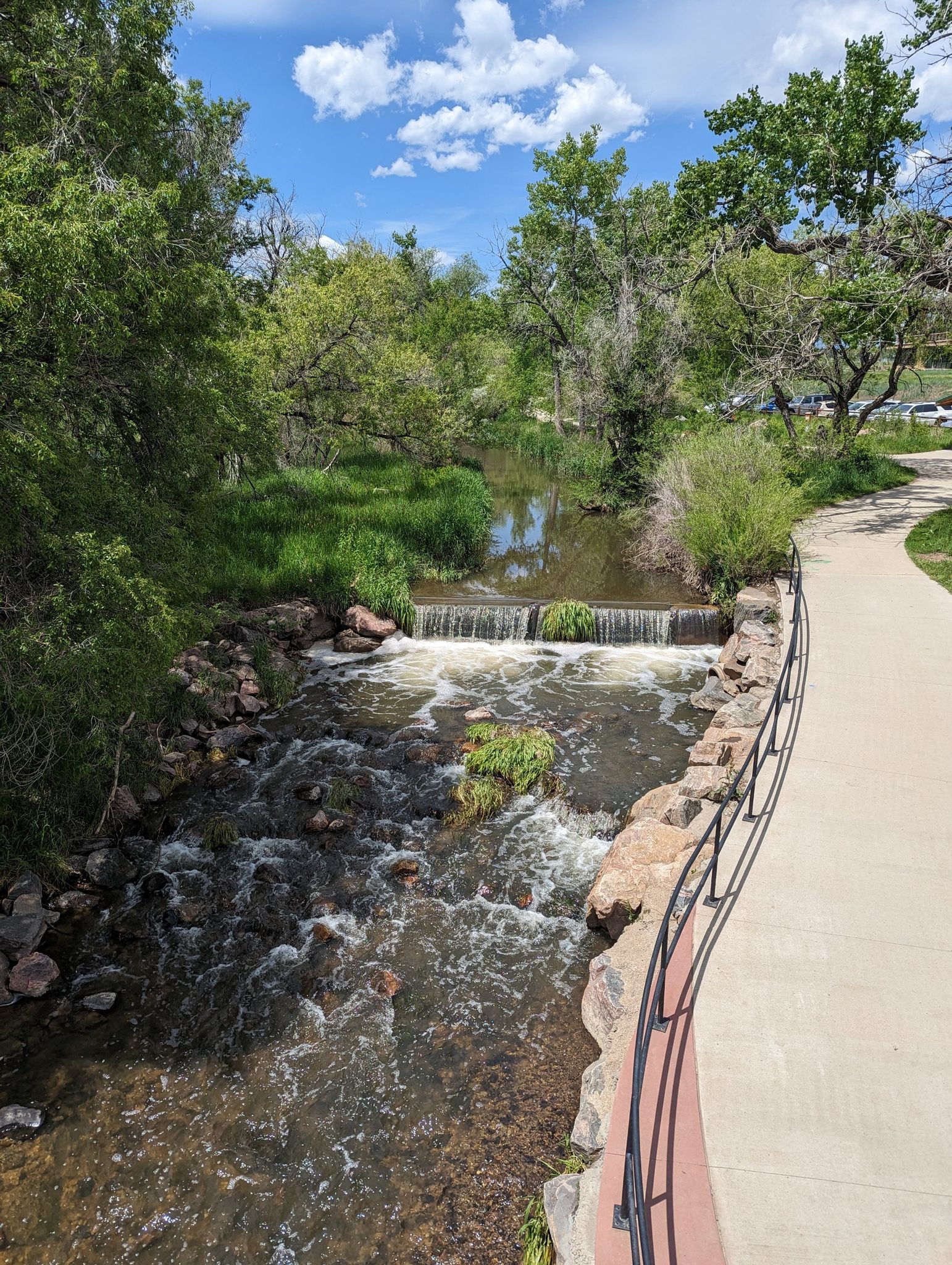 A flowing stream with small waterfalls next to a paved path and green trees. - Warembourg Open Space loop from Warembourg-Lafayette Farms
