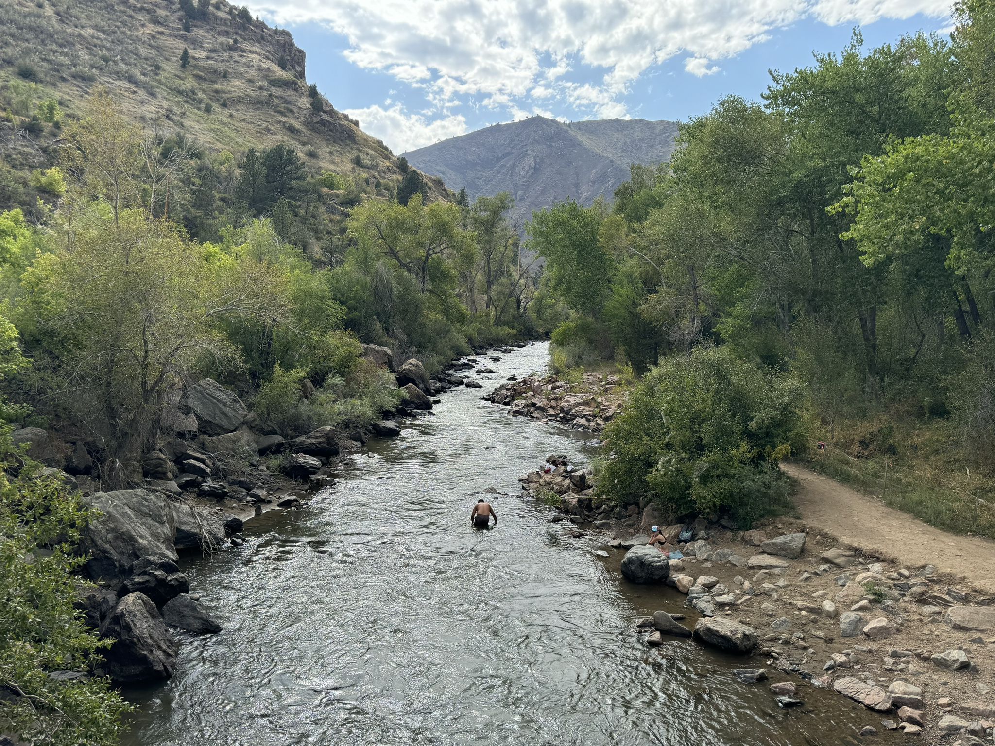 Man swimming in a river, surrounded by green trees and mountains under a cloudy sky. - Peaks to Plains and Welch Ditch Loop