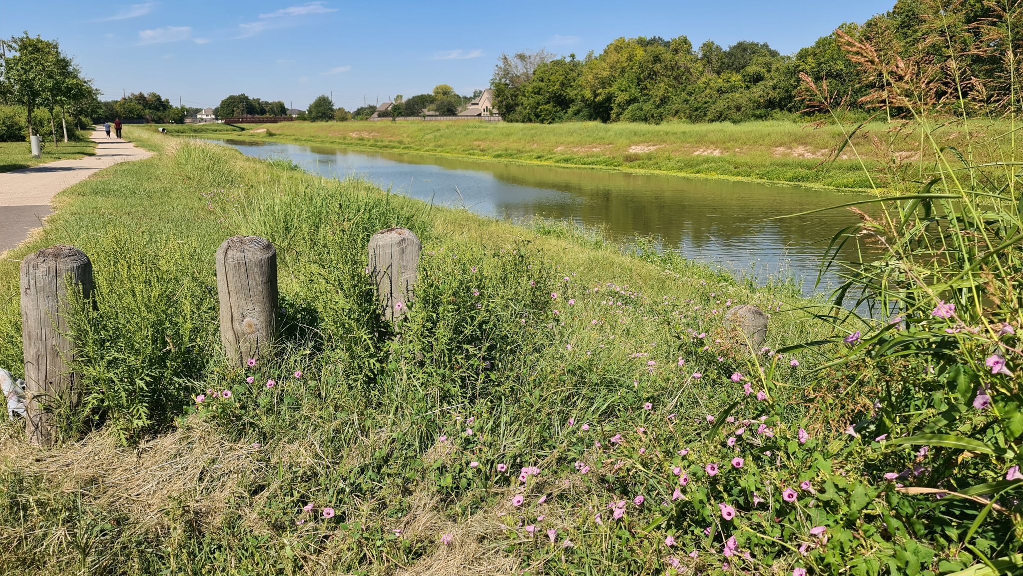 A narrow river flows between grassy banks with a path on a sunny day. - George Bush Hike & Bike Trail – George Bush Trail loop from Barker