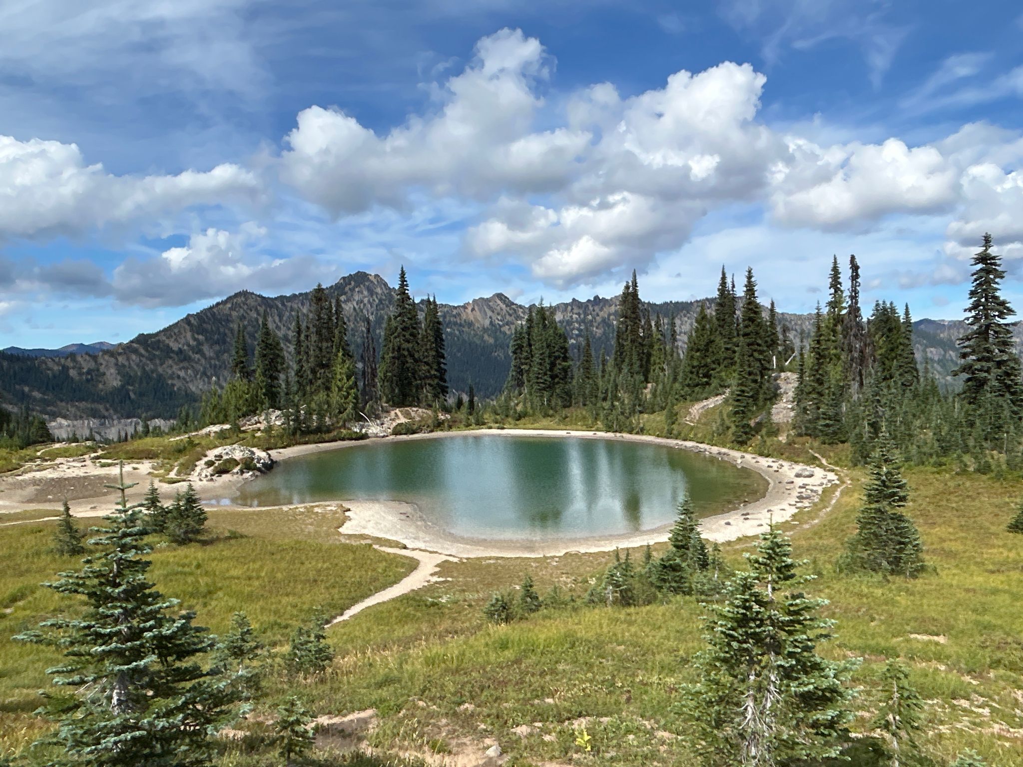 Serene mountain lake with reflections, surrounded by green trees and distant peaks. - Pacific Crest Trail: Section I - White Pass to Snoqualmie Pass