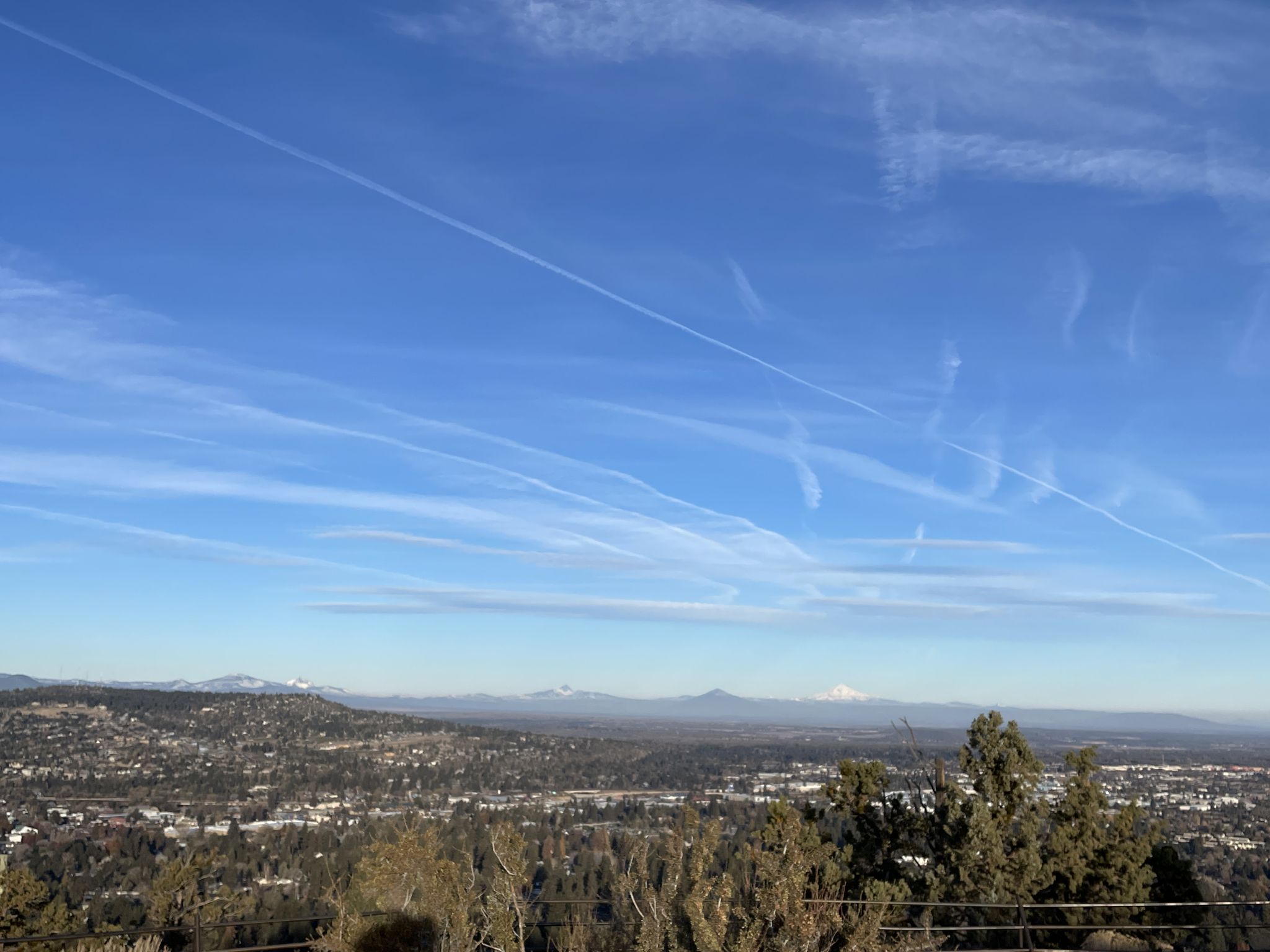 Cityscape with distant mountains under a clear blue sky with contrails. - Pilot Butte Nature Trail