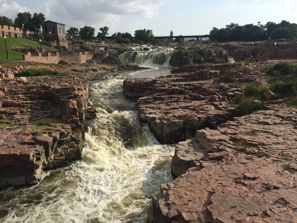 Rushing river water flows through a rocky gorge under a partly cloudy sky. - The Arc of Dreams – Falls Park Waterfalls loop from Sioux Falls Township