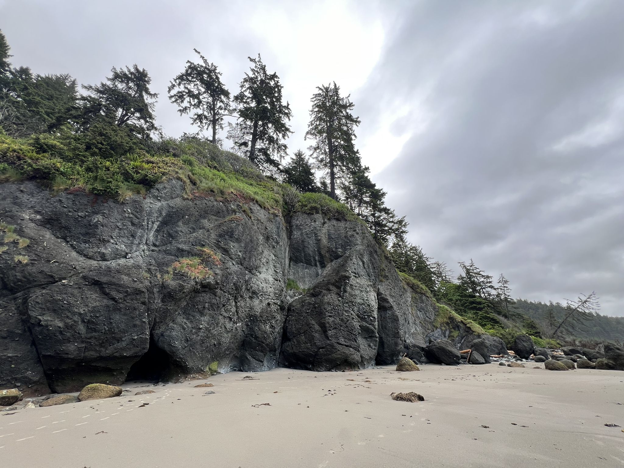 Overcast view of tall, dark rock cliffs rising from a sandy beach. - Hiking loop from Point of Arches State Park Property