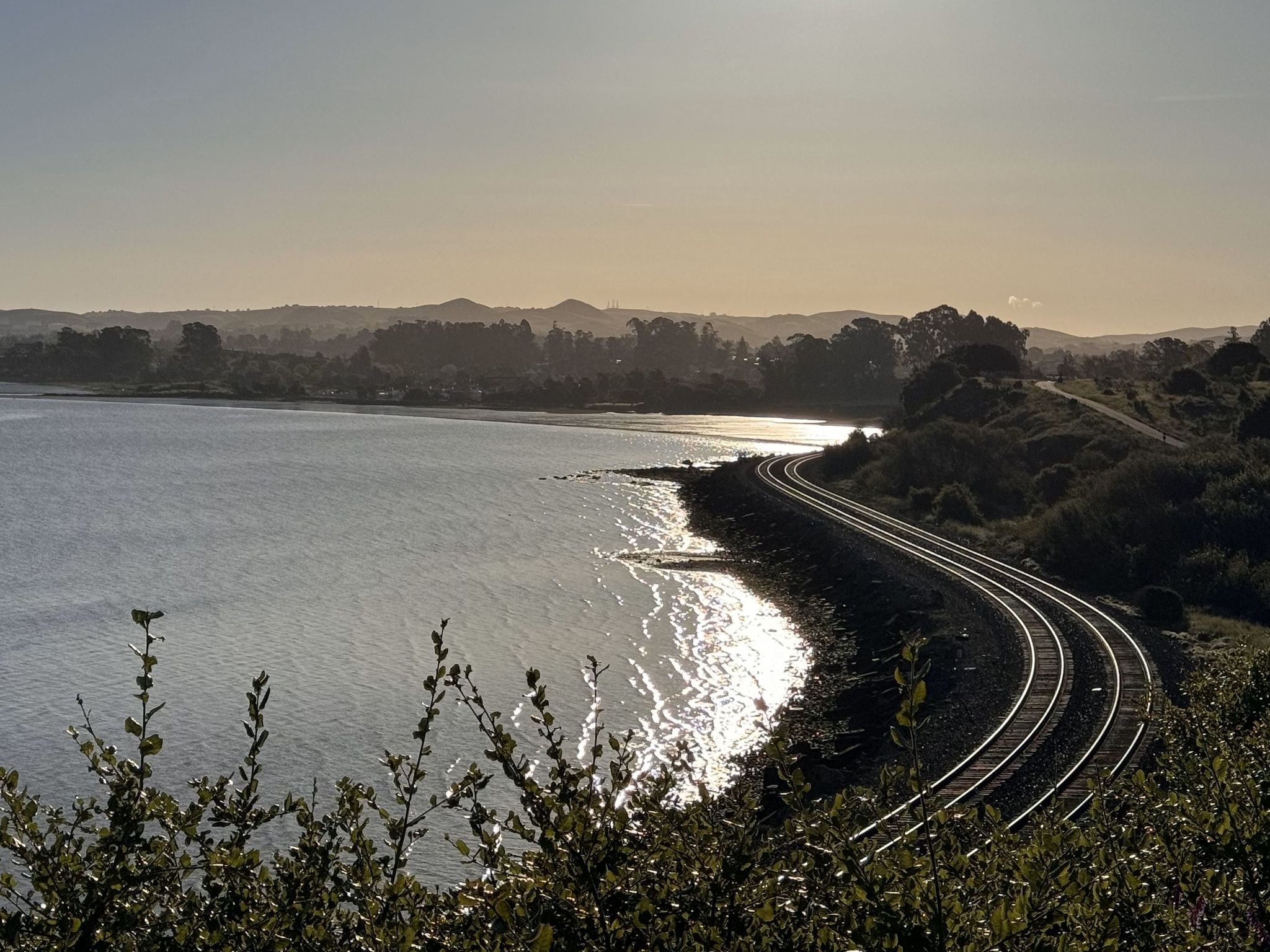 Golden hour light reflects on water next to curved railroad tracks and distant hills. - Pinole Shores