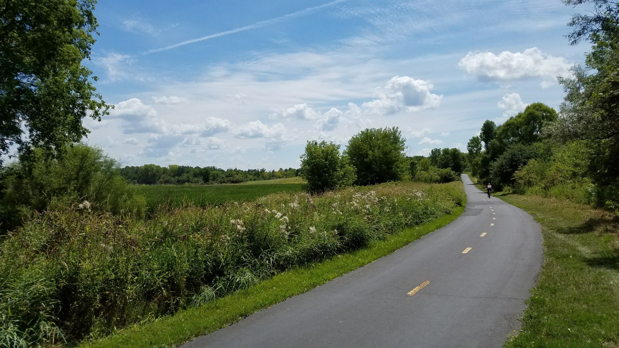 Paved path curving alongside a green field under a bright blue sky. - County road 50 – Kasma Marsh loop from County Road 110 Trail Access