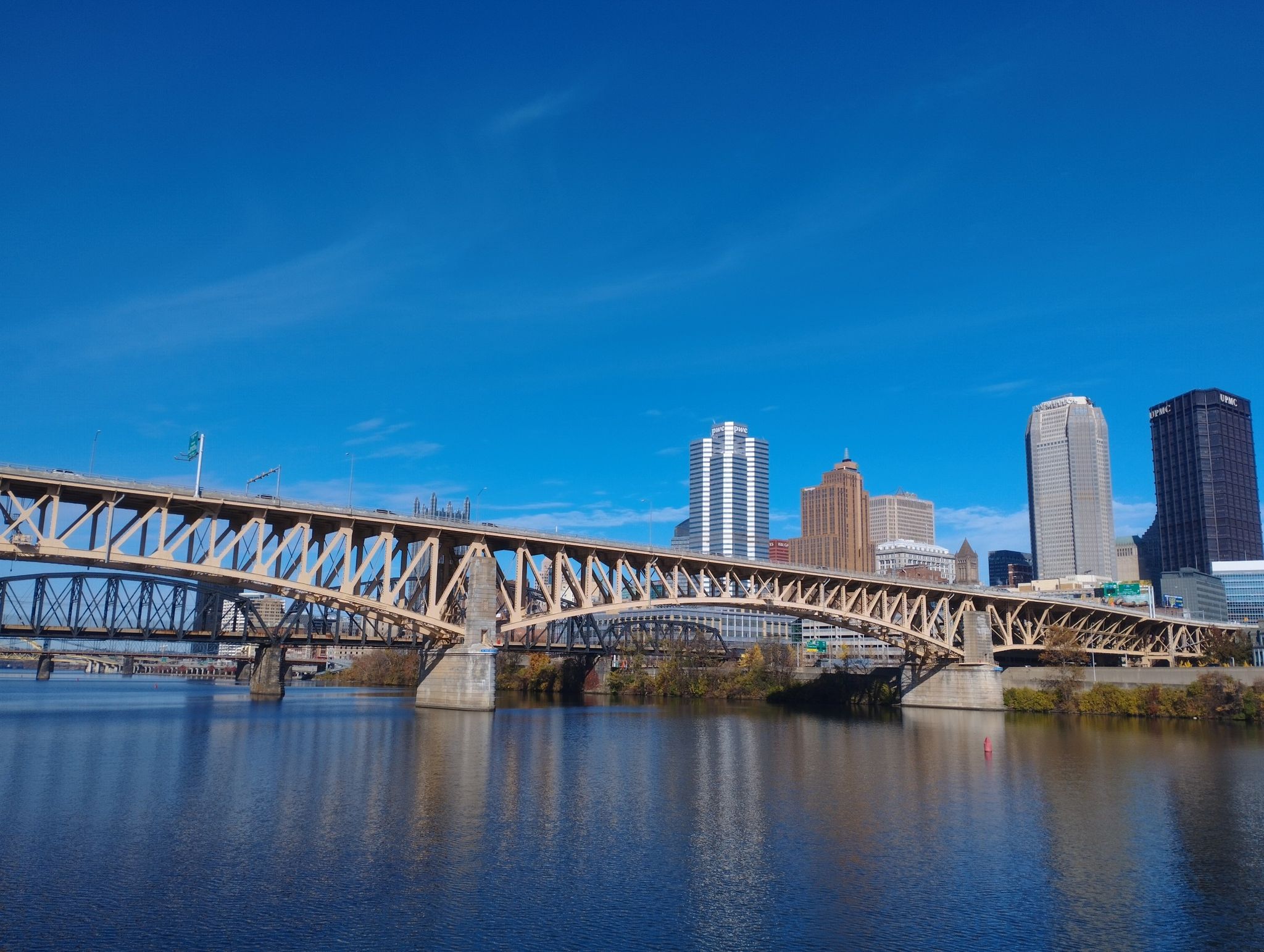 A large truss bridge spans a river with a city skyline under blue sky. - South Side Trail
