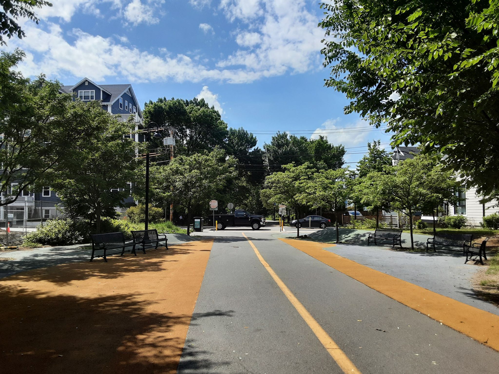 A paved path with yellow lines, trees, benches, and distant buildings. - Mass Central Rail Trail – Black Lives Matter graffiti loop from Somerville