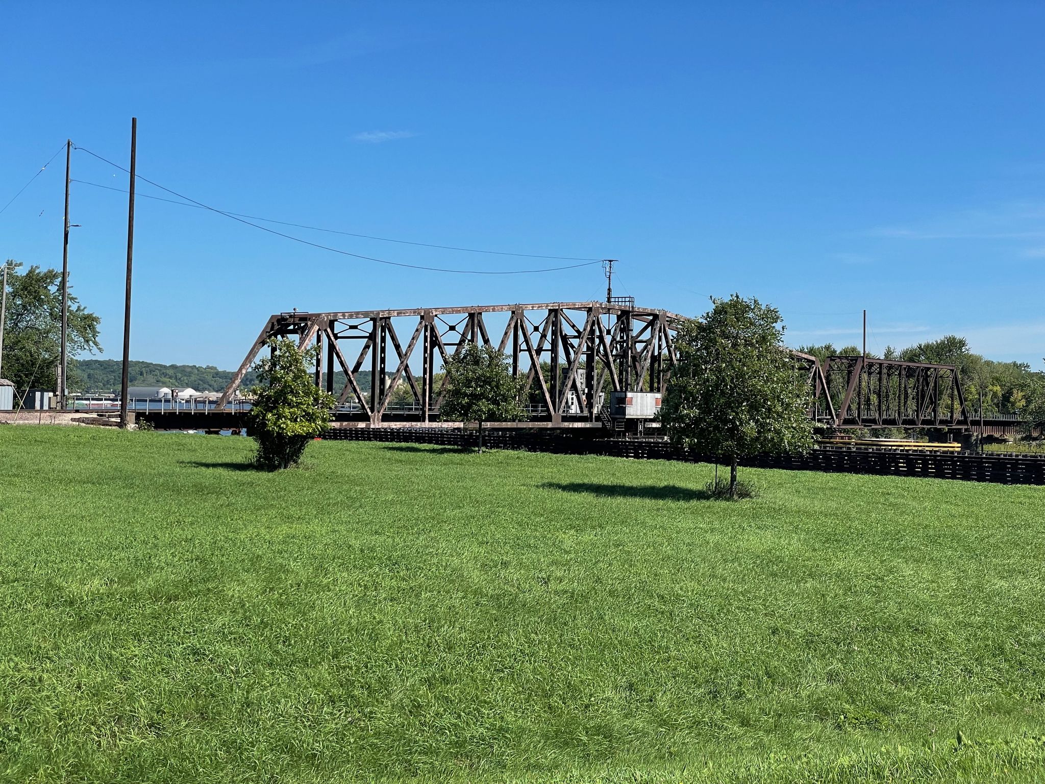 A metal truss bridge stands behind a vibrant green field under a clear blue sky. - Mississippi River Greenway