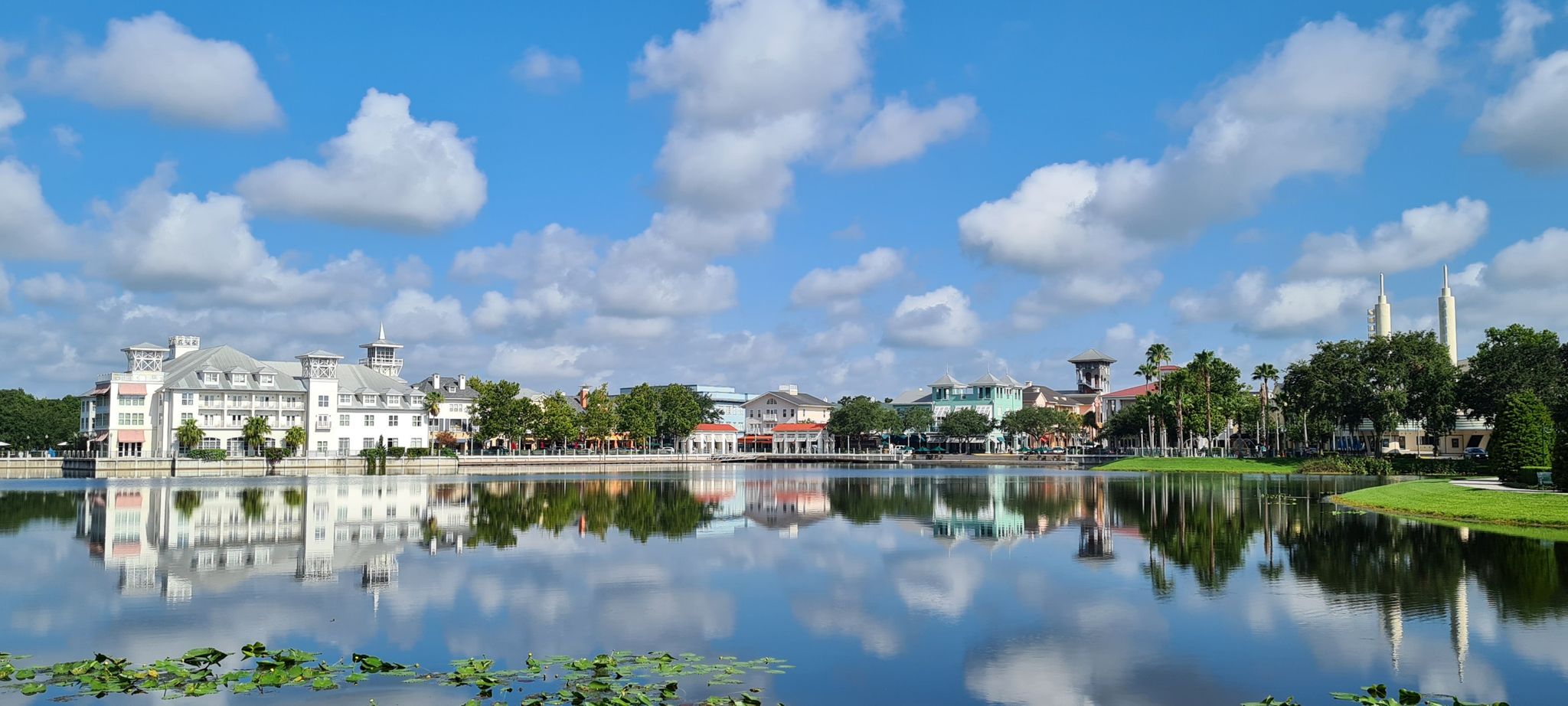 Buildings reflecting in a calm lake under a bright, cloudy blue sky. - Celebration Village loop via Lake Rianhar & Lake Evalyn