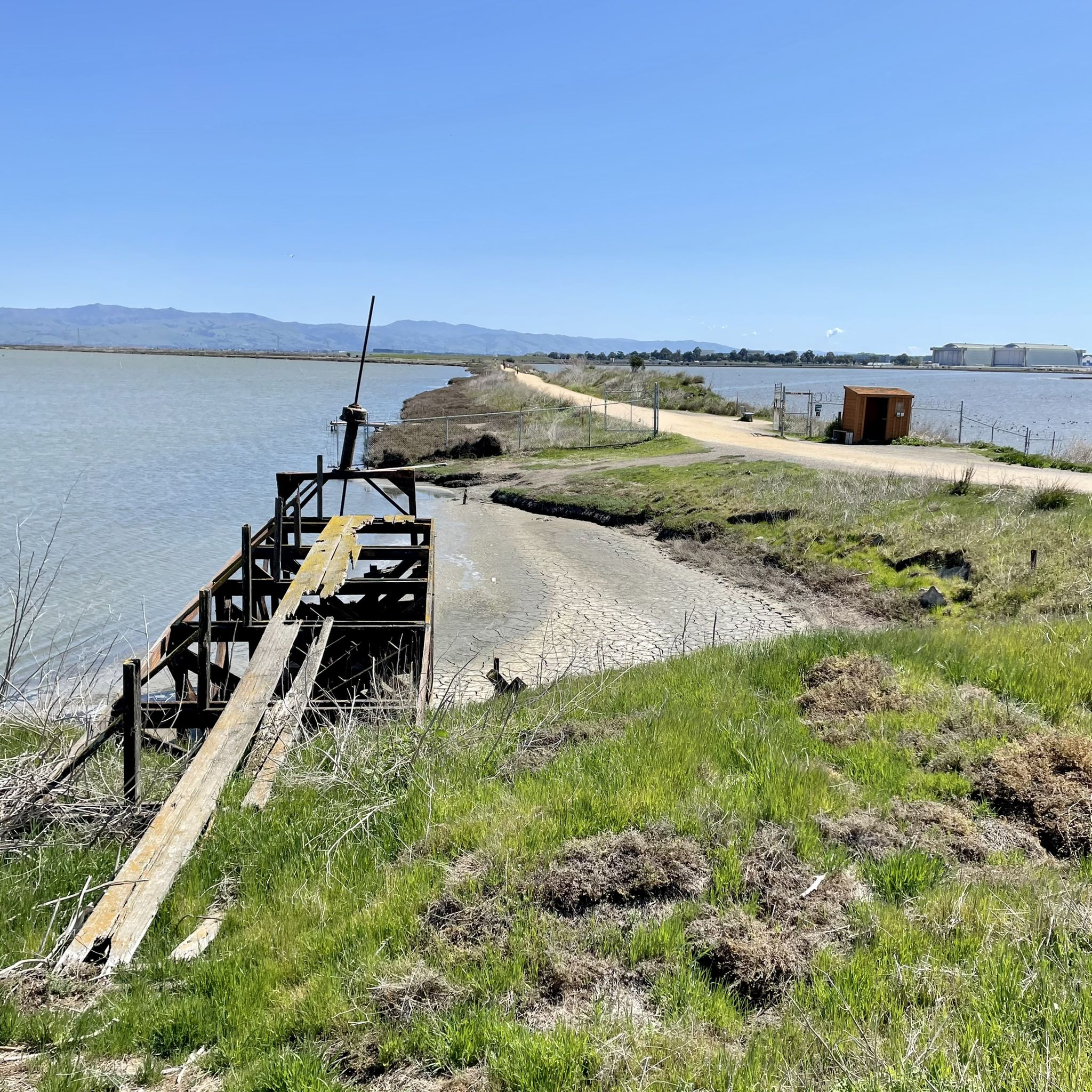 Wooden structure by calm water, dirt path, green grass, bright daylight. - Stevens Creek Trail