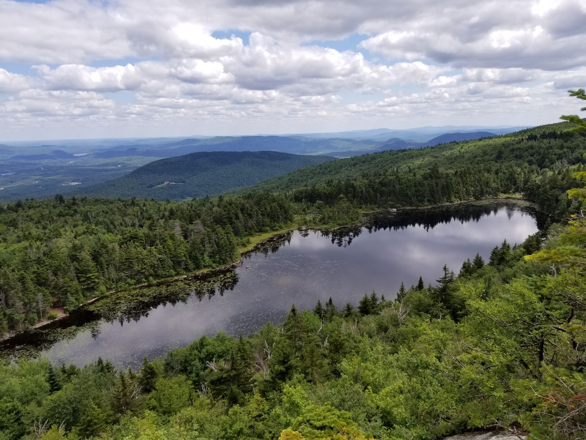Aerial view of a dark lake surrounded by lush green forest under a cloudy sky. - Mount Sunapee via Newbury Trail