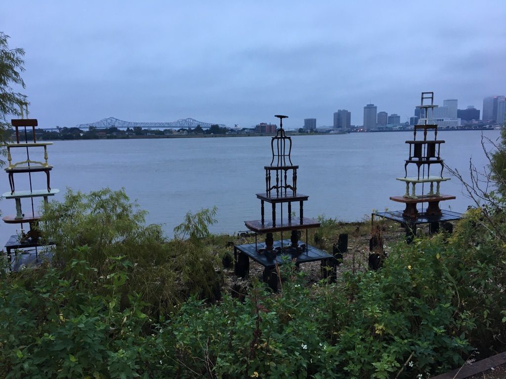 Three stacked furniture sculptures on a riverbank with a city skyline under an overcast sky. - Washington Artillery Park – Audubon Park loop from French Quarter