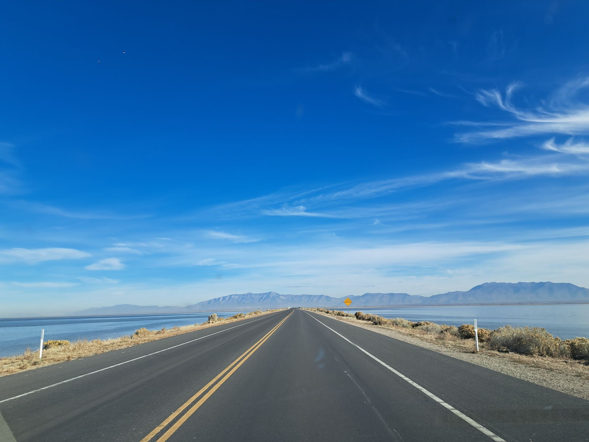 A long, straight road stretches between water and distant mountains under blue sky. - Bear Sighting loop from Syracuse Utah West Stake