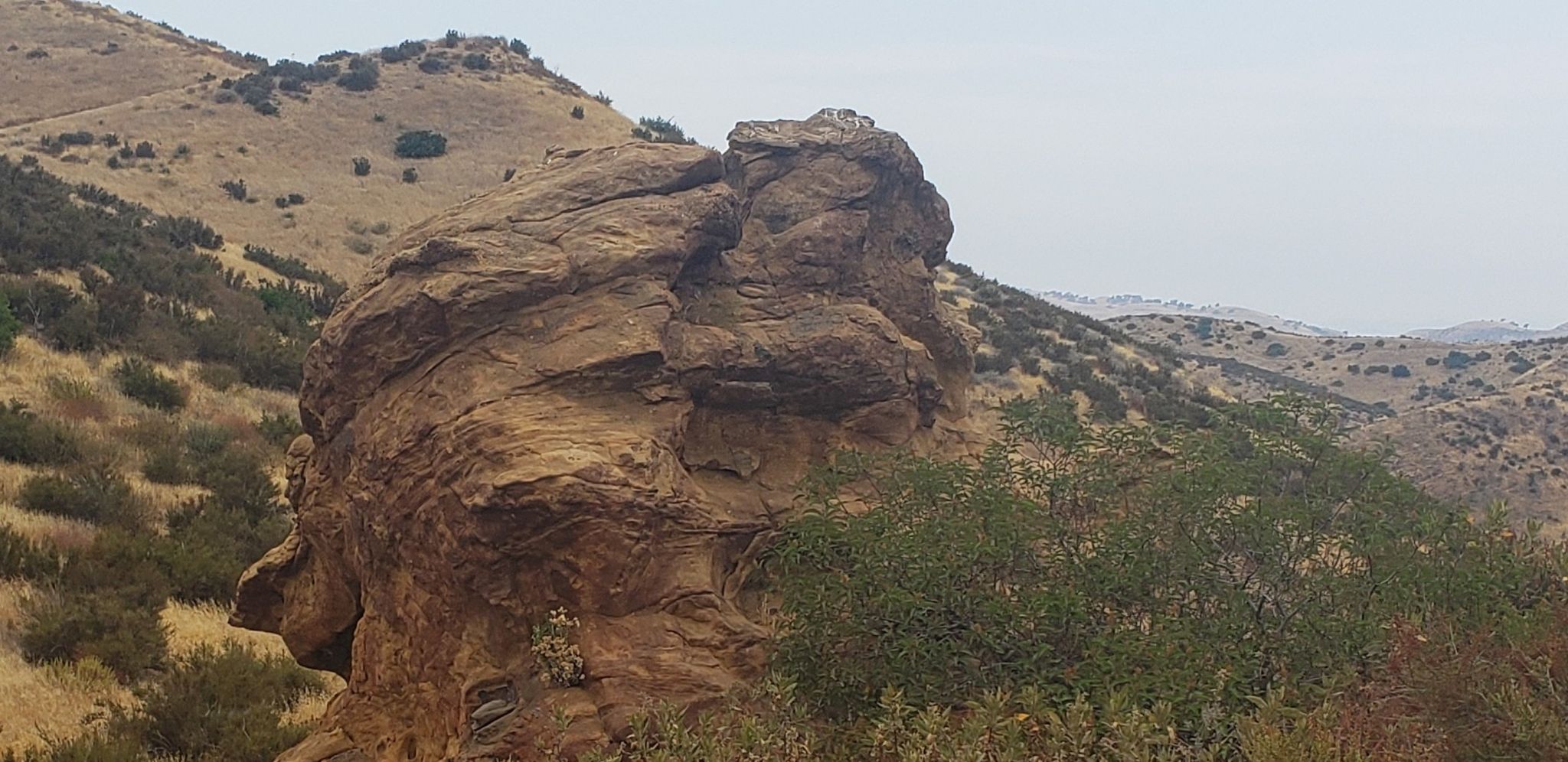 Large, weathered rock formation stands among dry, brushy hills. - Las Llajas Canyon Trail