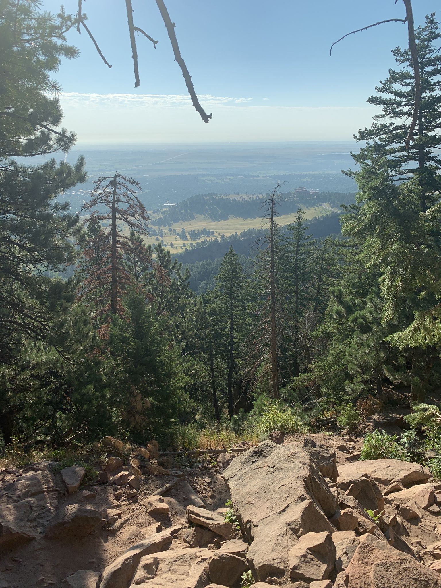 A rocky foreground and pine trees overlook a vast, hazy valley and distant plains. - Royal Arch Trail