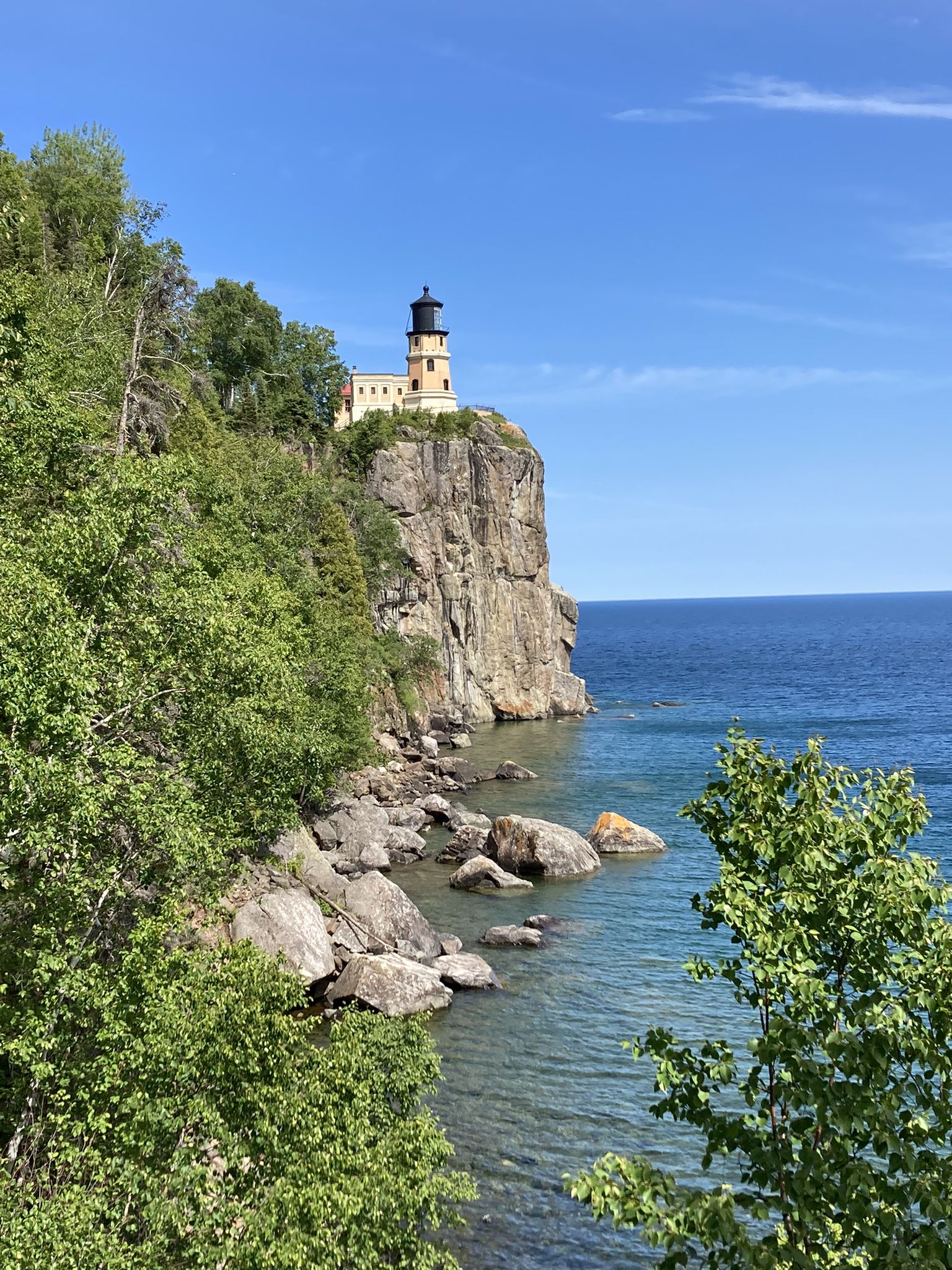 Lighthouse on a rocky cliff above clear blue water under a sunny sky. - Gitchi-Gami State Trail to Split Rock Lighthouse
