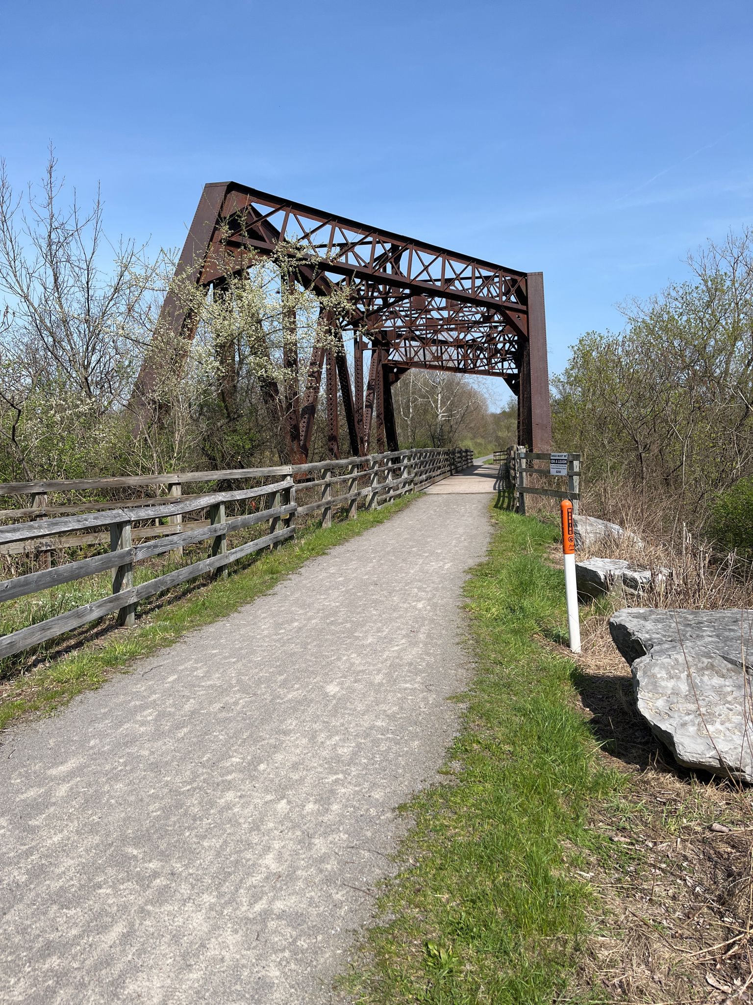 Gravel path leads to a large, rusty metal truss bridge under a clear blue sky. - Lehigh Valley Trail – Leigh Valley Trail loop from Honeoye Falls